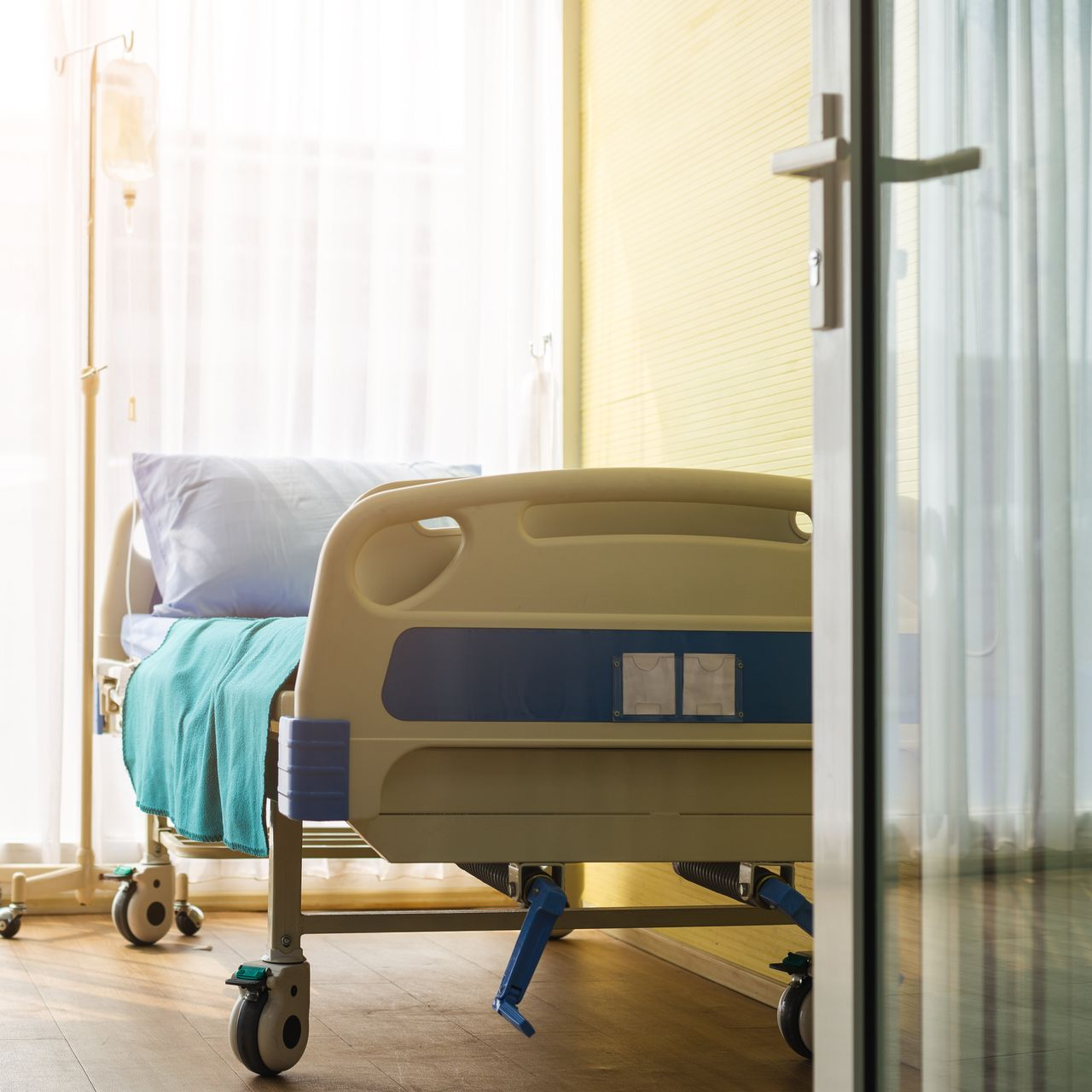 Lit d'hôpital près d'une fenêtre lumineuse avec des rideaux blancs et une couverture bleue dans une chambre.