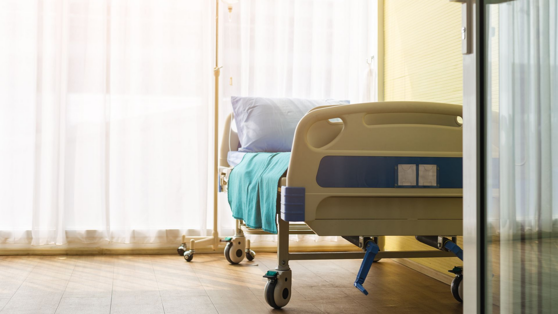 Lit d'hôpital jaune près d'une fenêtre lumineuse, avec des rideaux blancs et une couverture turquoise.