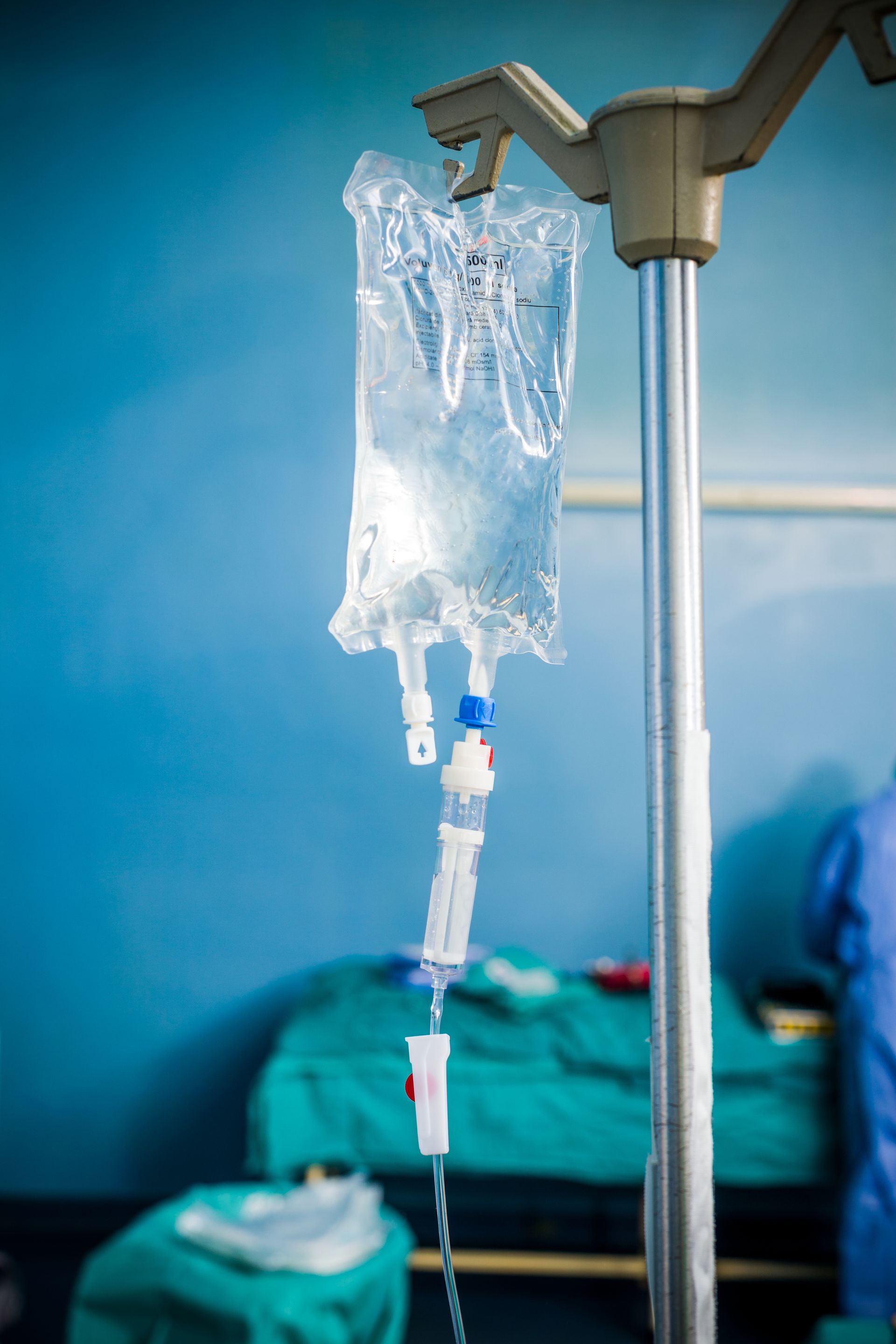 Poche de perfusion intraveineuse suspendue à un pied d'hôpital près d'un lit aux draps bleus et verts.