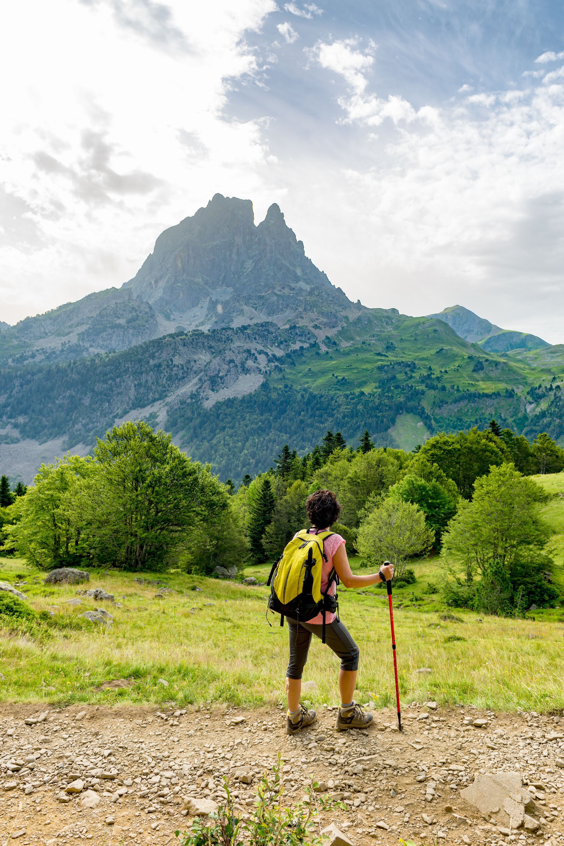 Randonneur avec un sac à dos jaune sur un sentier rocailleux face à une montagne escarpée sous un ciel nuageux et lumineux.