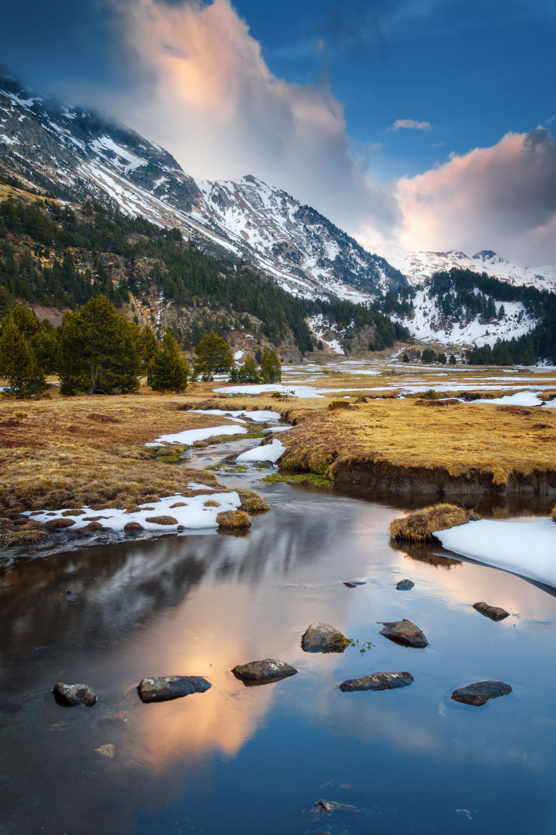 Montagne enneigée se reflétant dans un ruisseau calme au bord d'une vallée herbeuse au coucher du soleil.