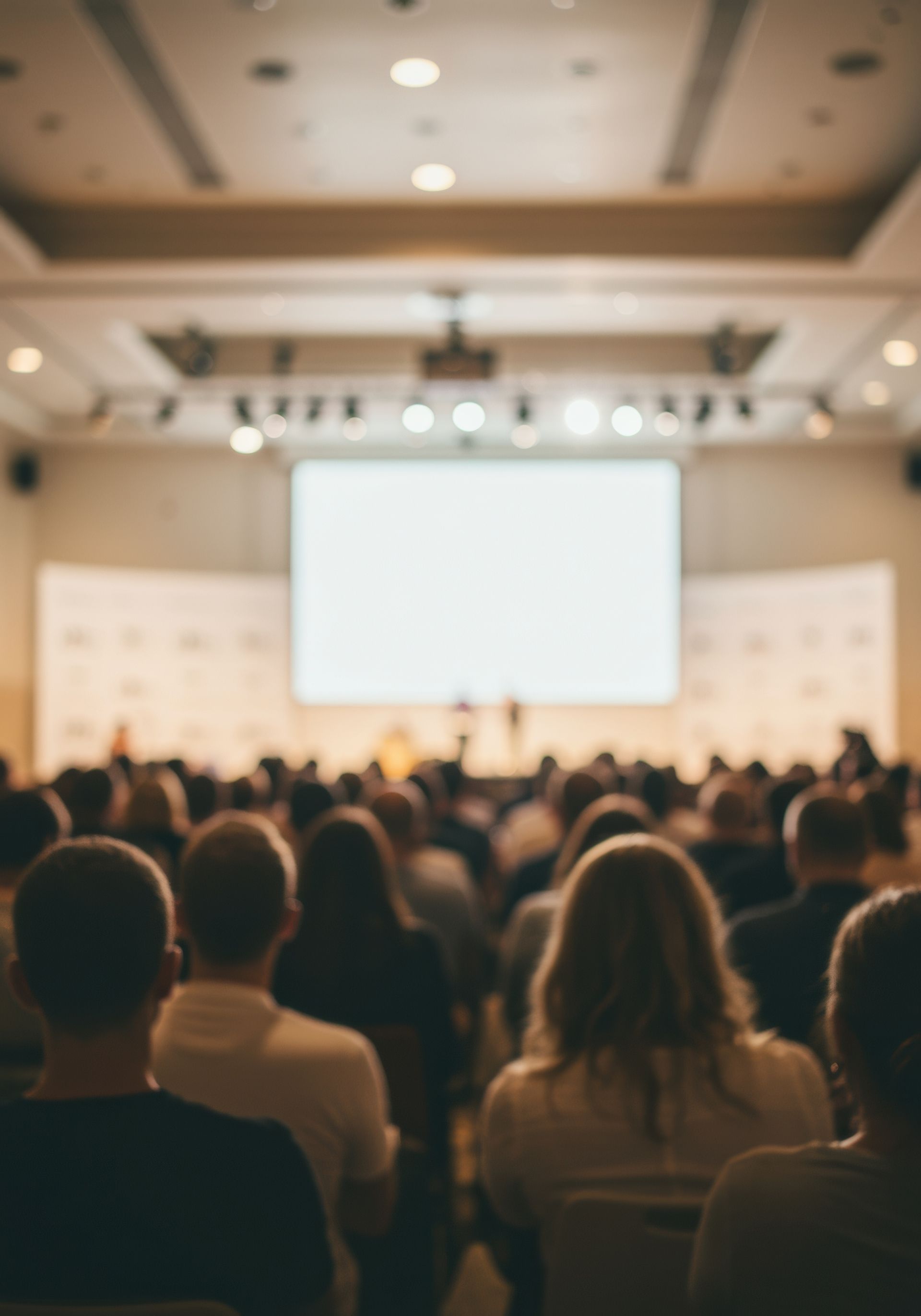 Le public est assis dans une salle de conférence, face à un écran de projection lumineux sur scène.