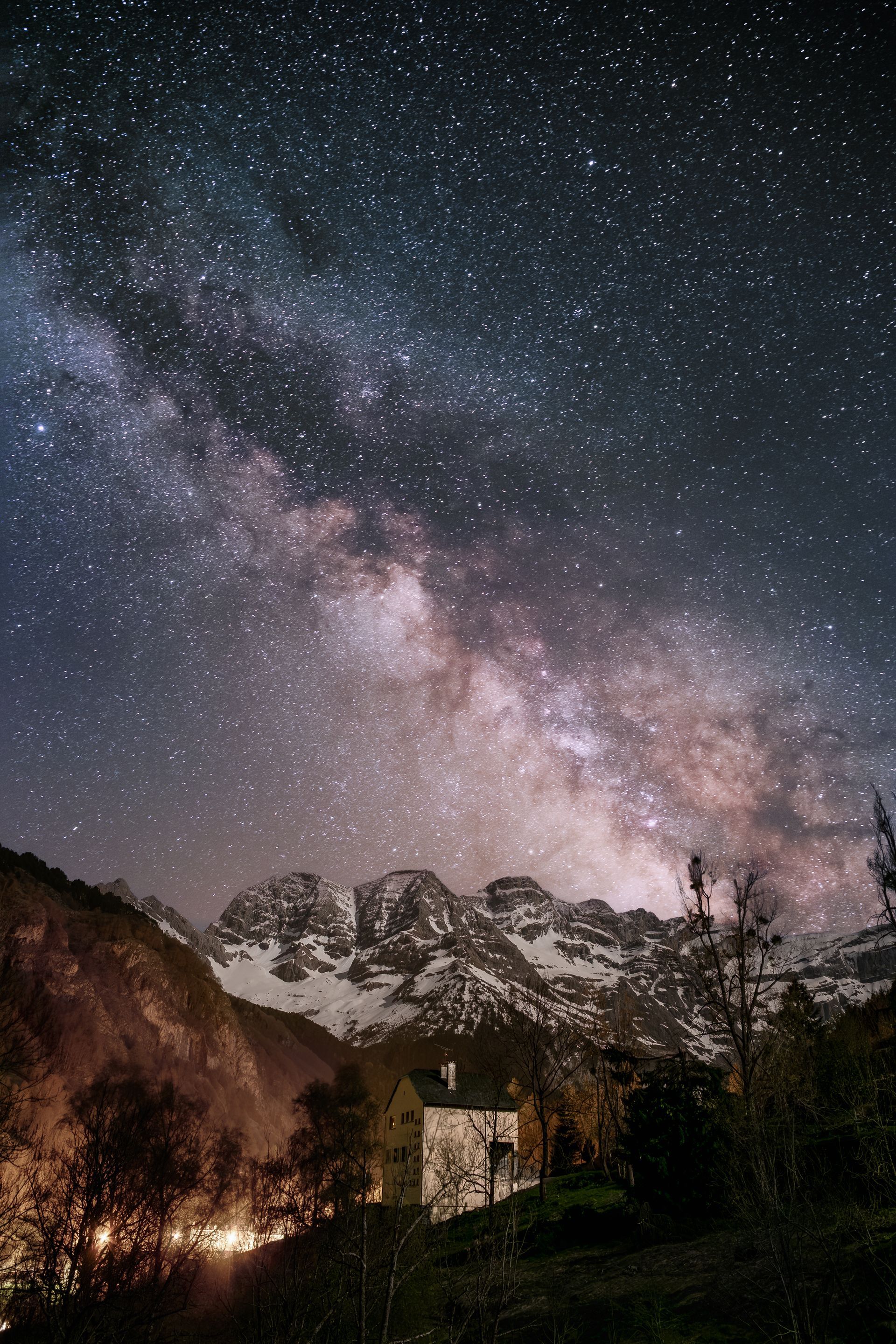Voie lactée au-dessus d'un chalet de montagne sombre avec des arbres en silhouette la nuit.