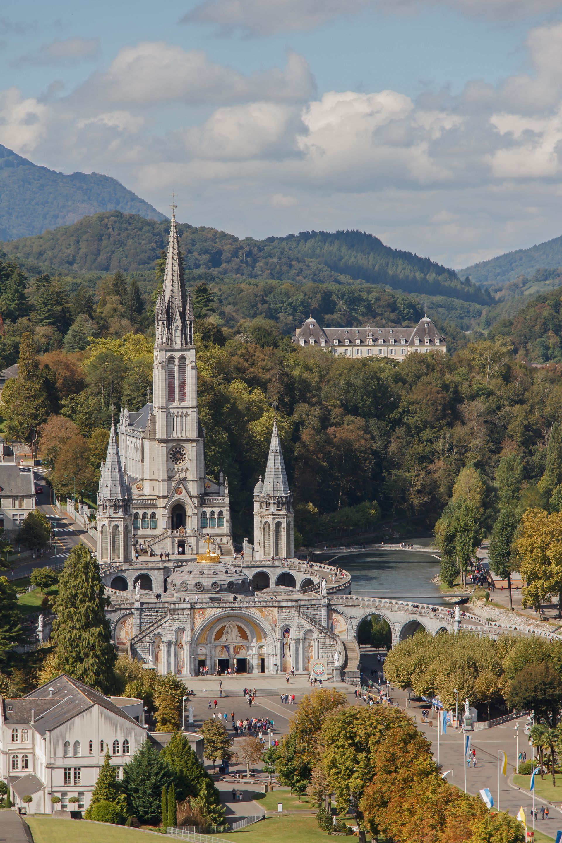 Église ornée avec une haute flèche dans une vallée boisée, vue d'en haut sous un ciel nuageux.