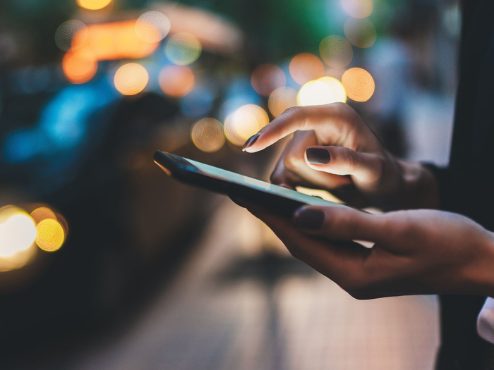 Des mains utilisent un smartphone la nuit, avec des lumières de la ville floues en arrière-plan.