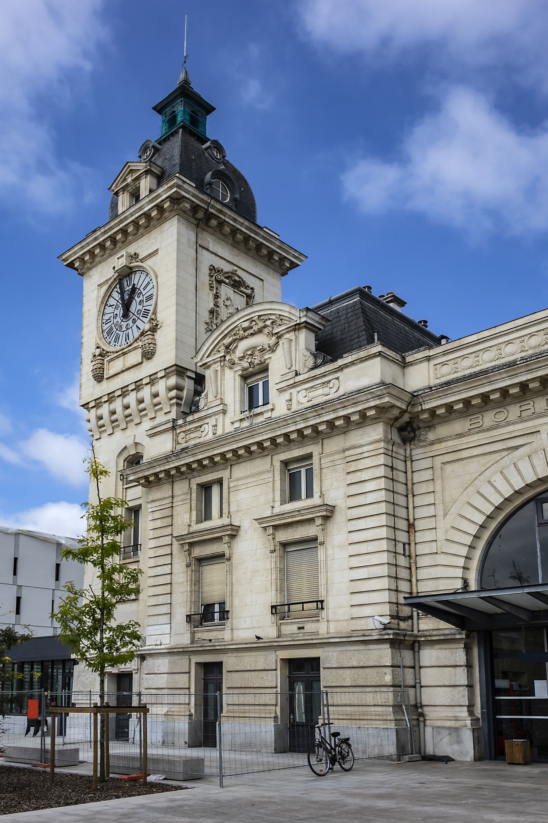 Bâtiment historique en pierre avec tour d'horloge sous un ciel bleu, vu de la rue.