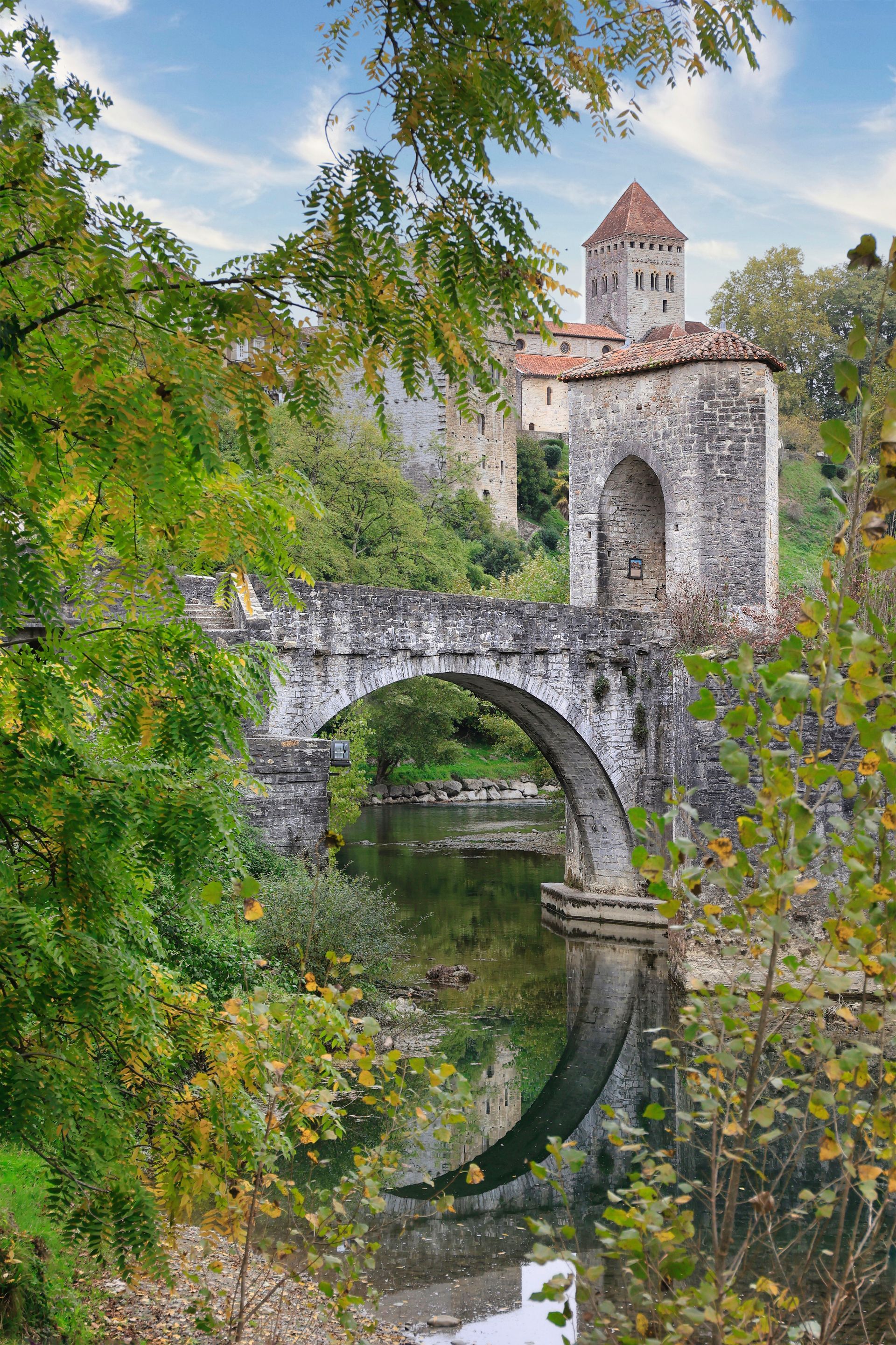 Pont de pierre et tour arquée enjambant une rivière calme, encadrés par une végétation luxuriante et ses reflets.