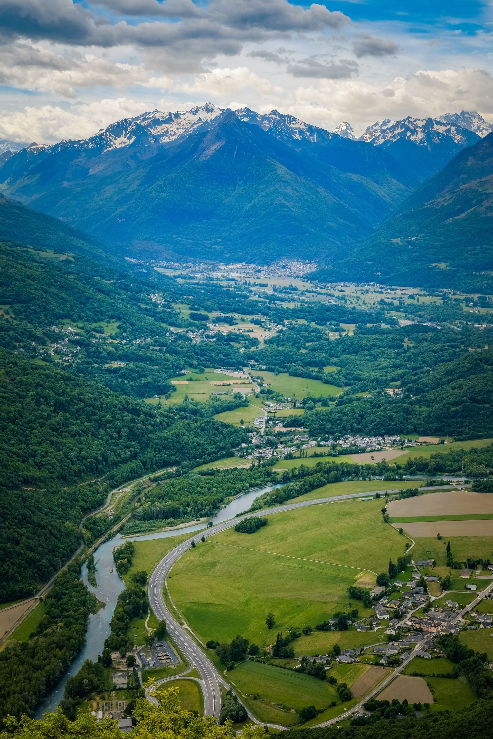 Vallée de montagne traversée par une rivière sinueuse, des champs verdoyants et des sommets enneigés sous un ciel nuageux.