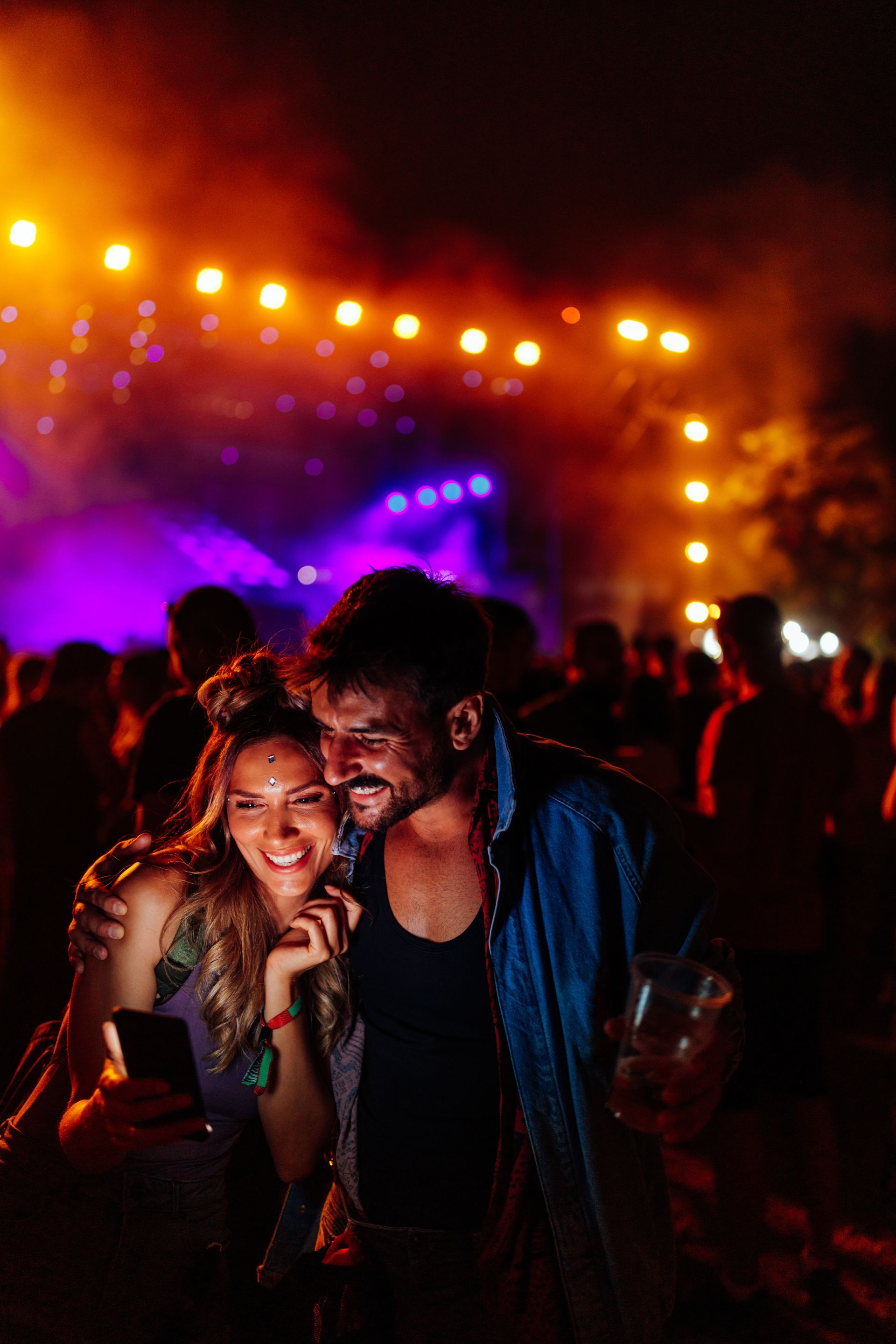 Un couple souriant à un concert, éclairé par des lumières de scène violettes et orange dans une salle bondée.