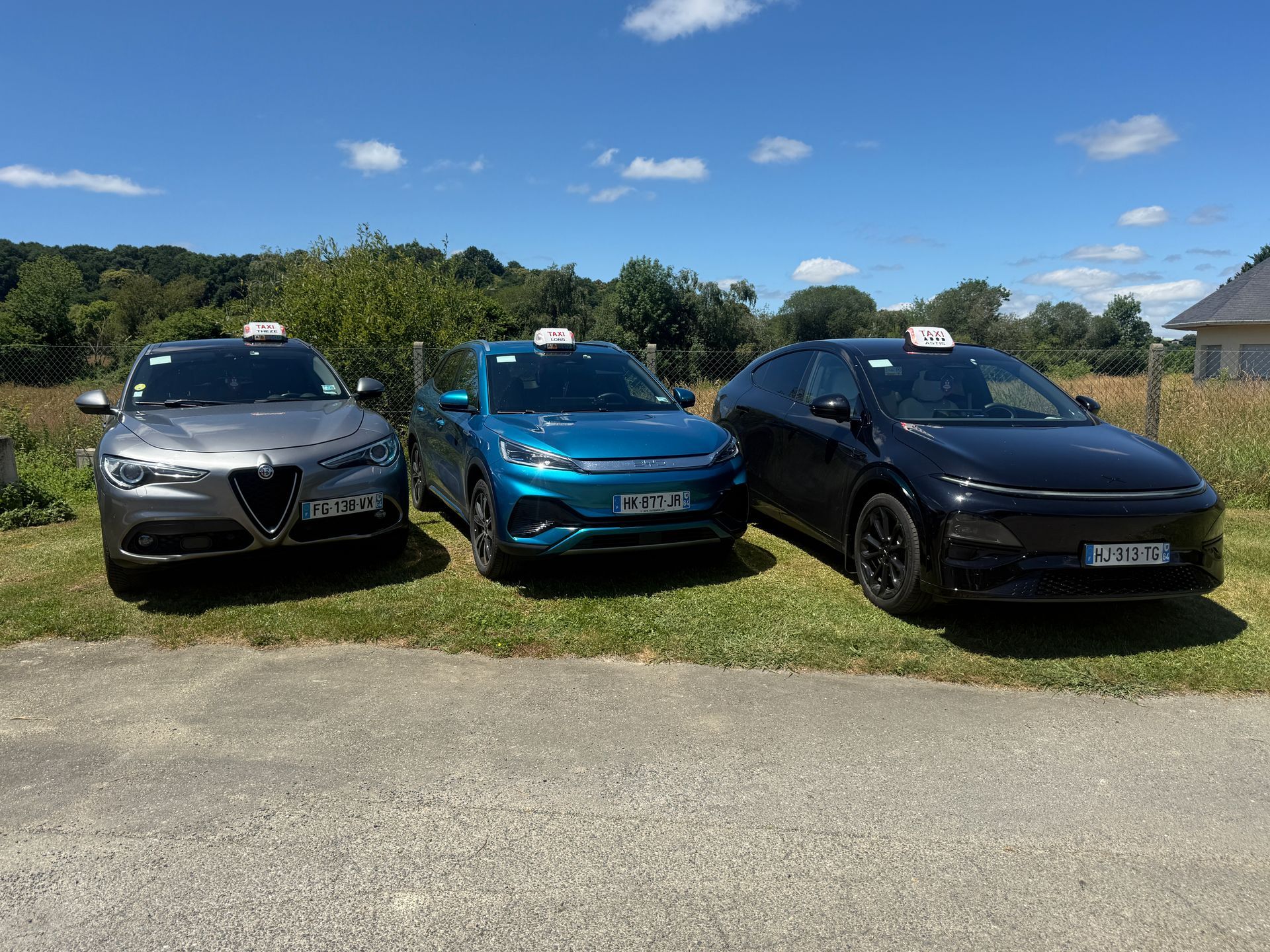 Trois voitures garées sur l'herbe sous un ciel bleu, dont une berline argentée, une voiture à hayon turquoise et un SUV noir.