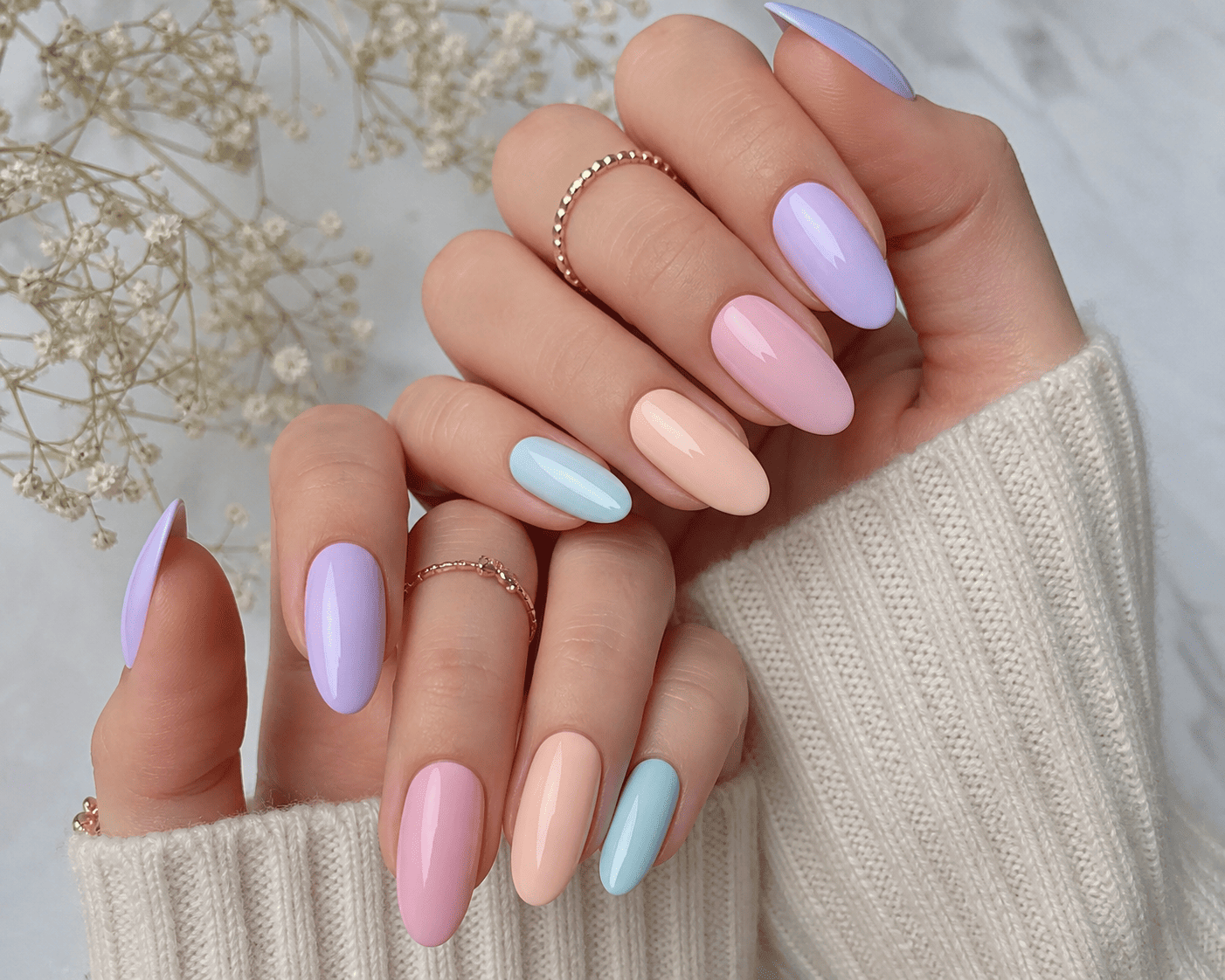 Des mains aux ongles manucurés dans des tons pastel lavande, rose et bleu, sous un pull crème.
