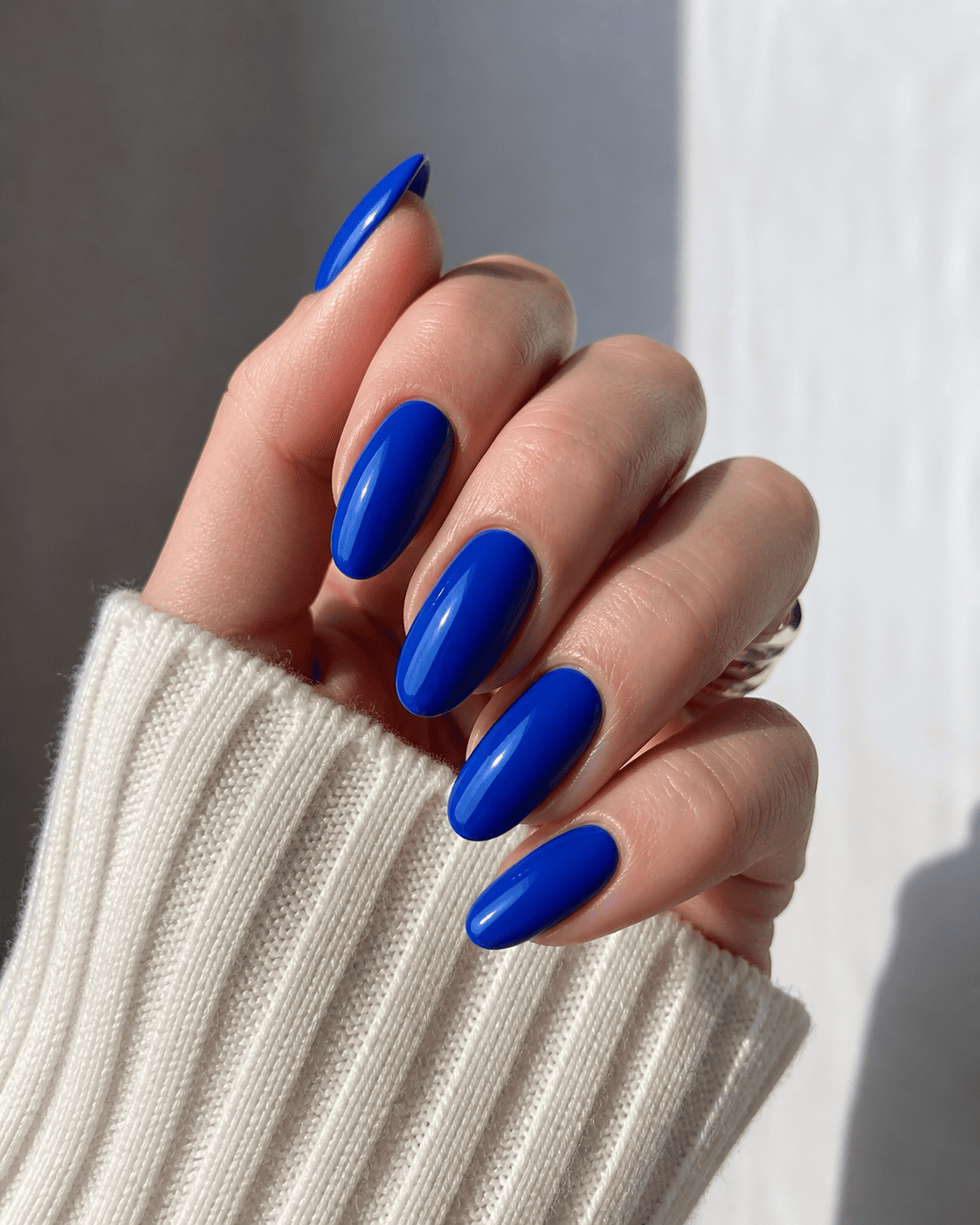 Main aux longs ongles bleu cobalt brillants, contrastant avec un pull côtelé crème.