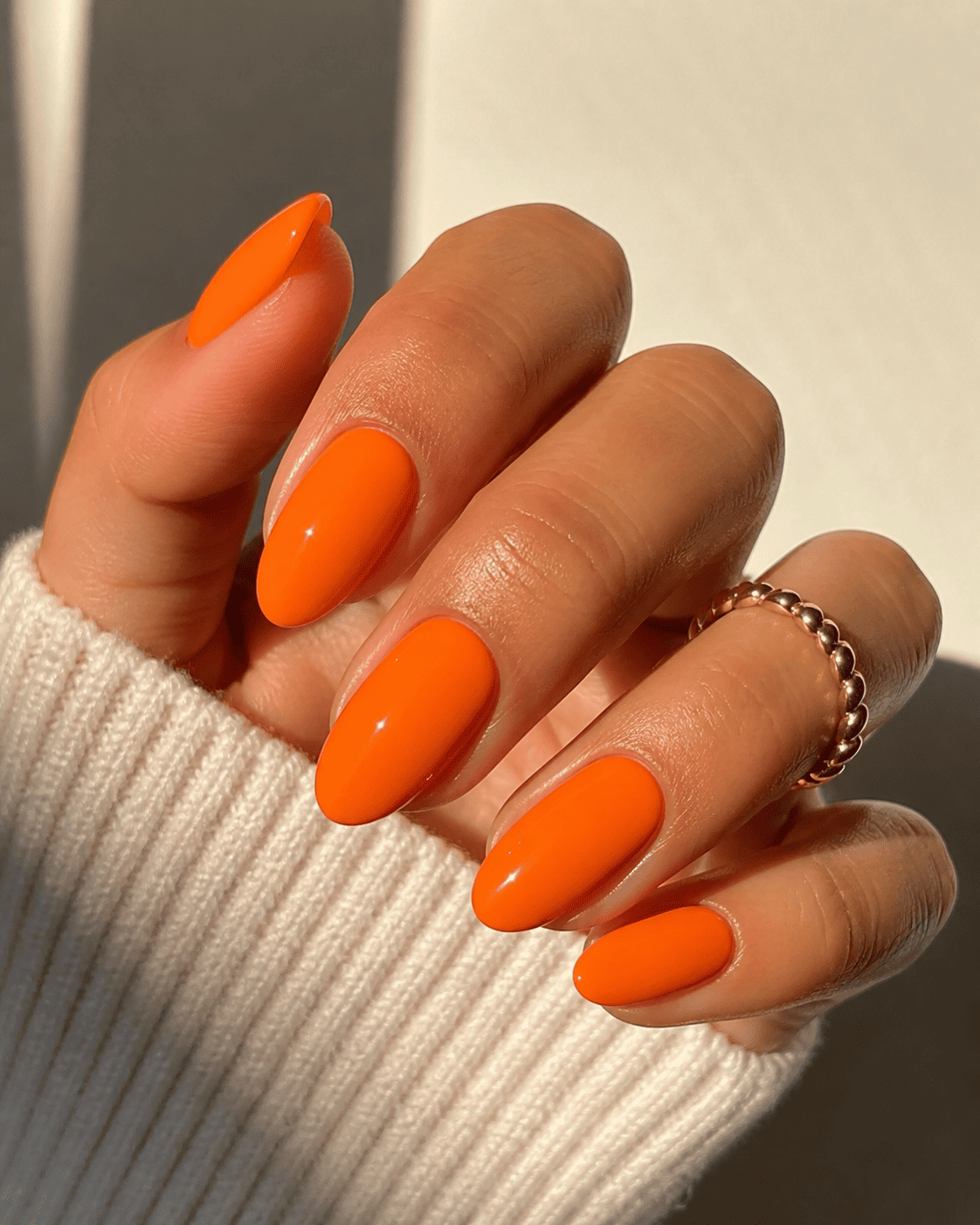 Main aux ongles orange vif ornée d'une bague en or, posée sur un pull côtelé crème.