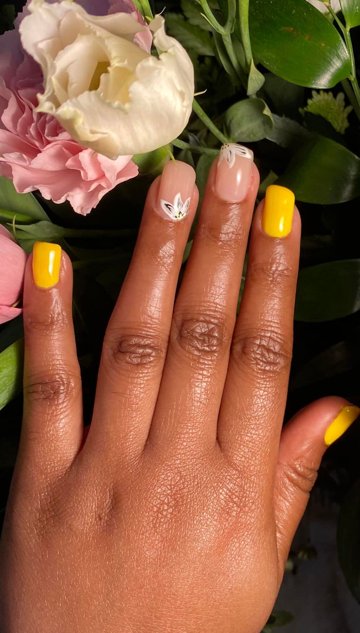 Main aux ongles jaune vif et ornée d'un accent blanc sur un doigt, posée près de fleurs roses et blanches.