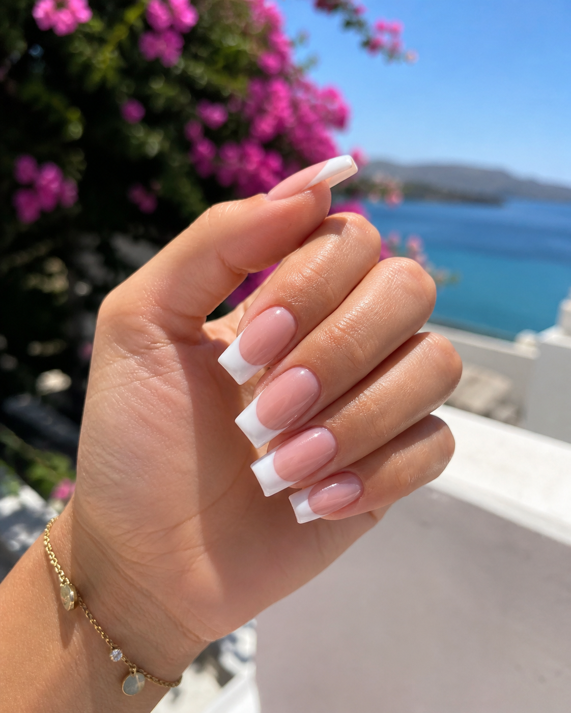 Main avec une manucure française rose et blanche tendue devant une vue ensoleillée sur la mer.