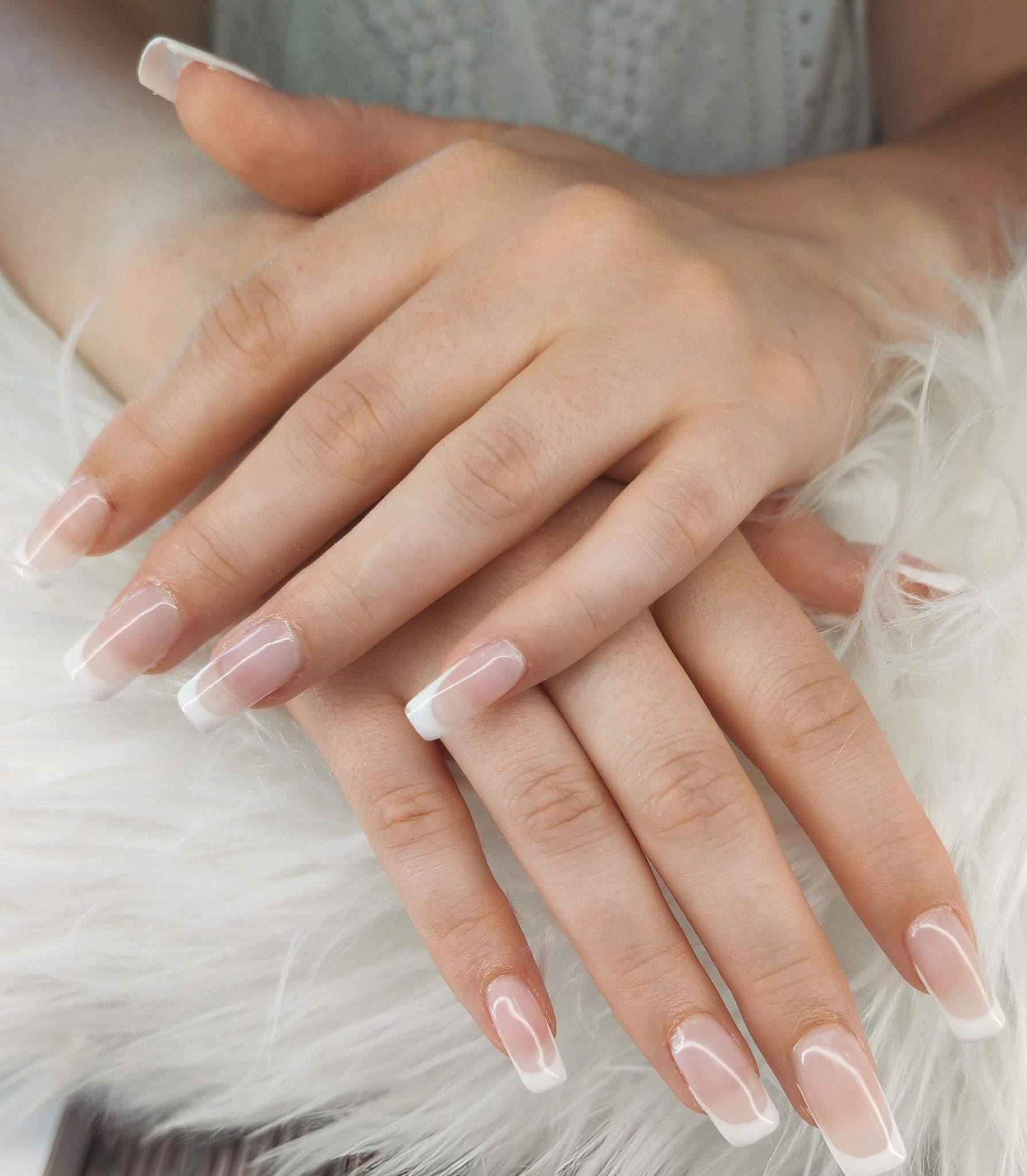 Des mains aux longs ongles rose pâle, vernis à la française, reposent sur une surface de fourrure blanche.