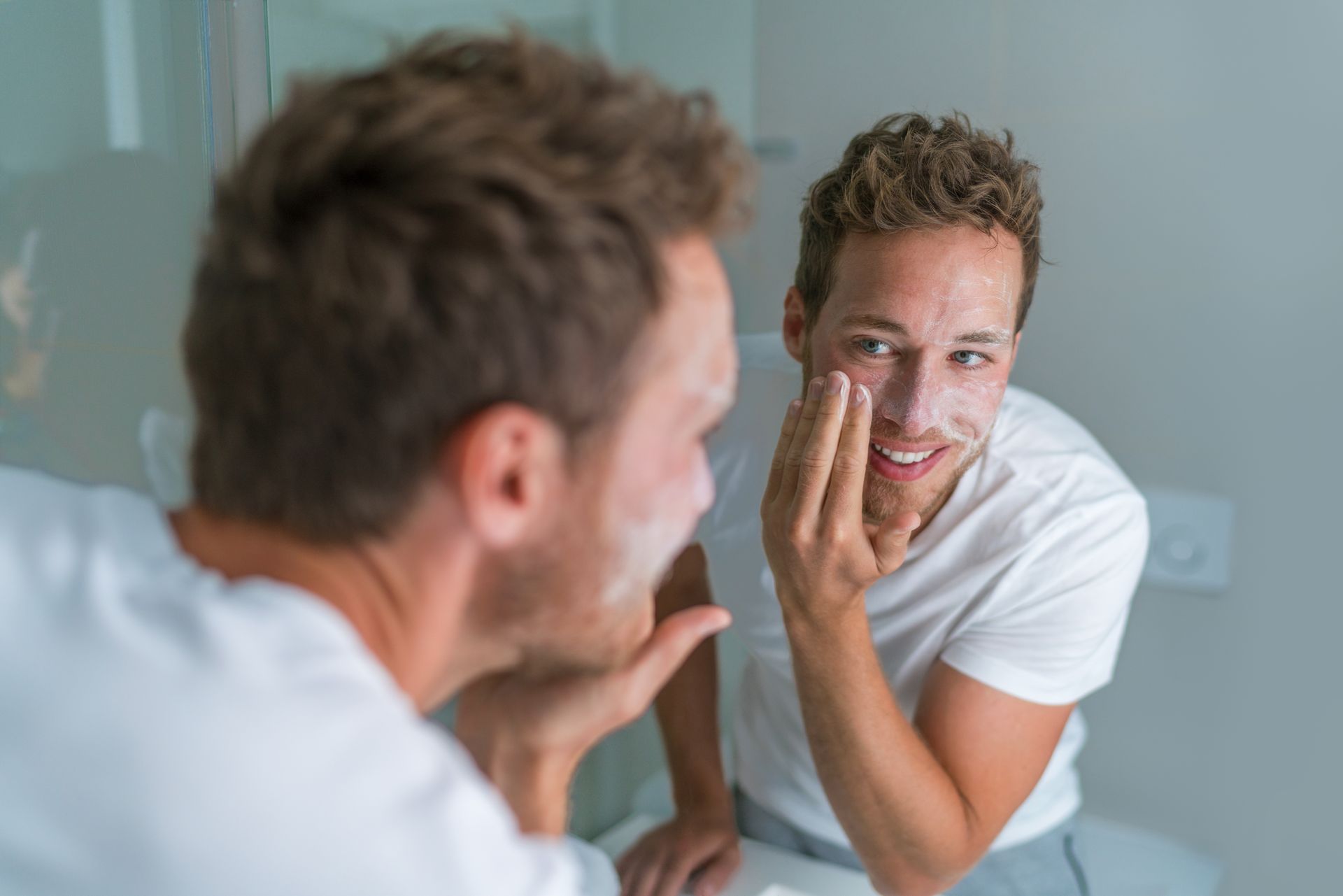Un homme souriant utilise un gommage pour le visage devant son miroir de salle de bains.