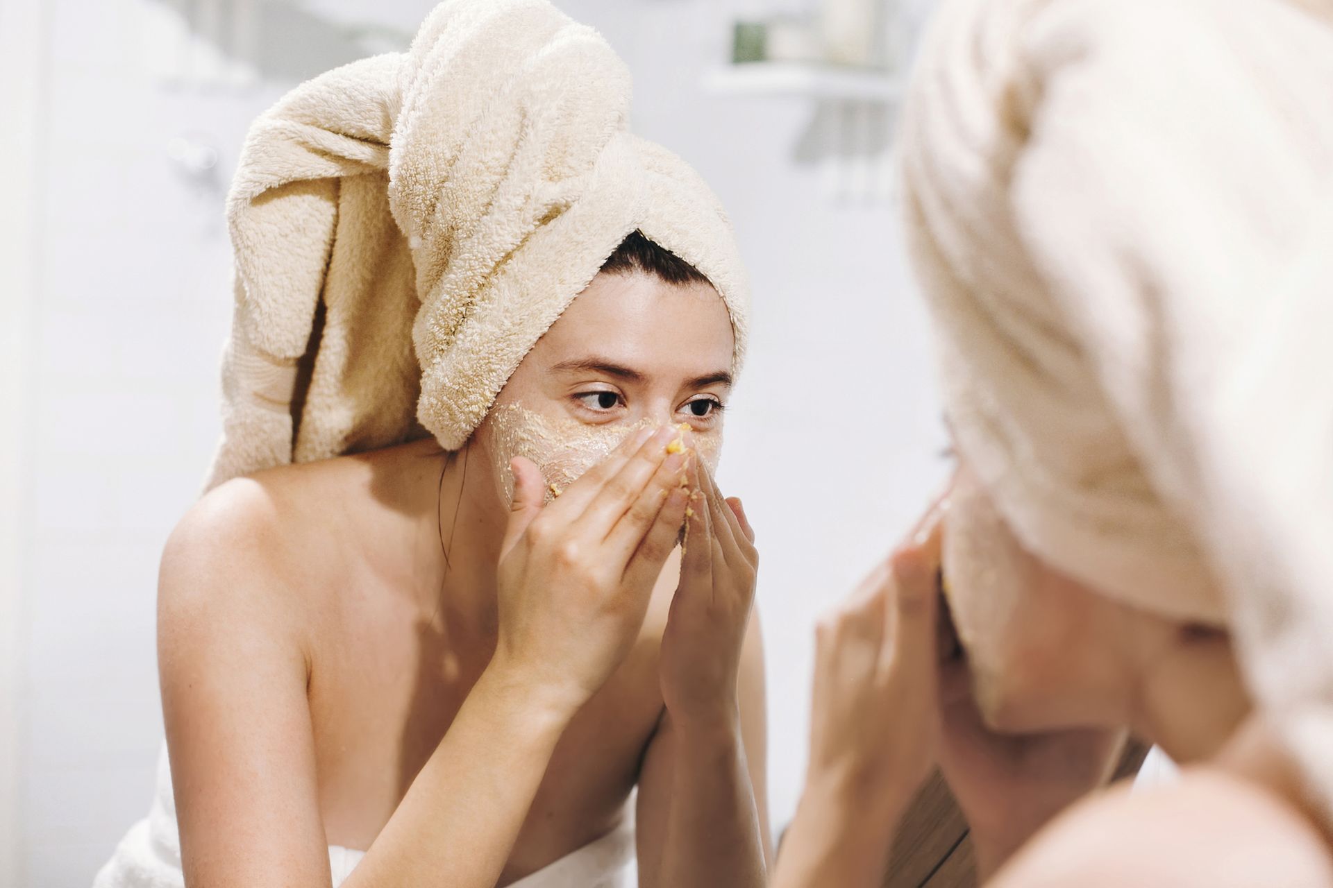 Une femme exfolie son visage avec un gommage en se regardant dans un miroir de salle de bains.