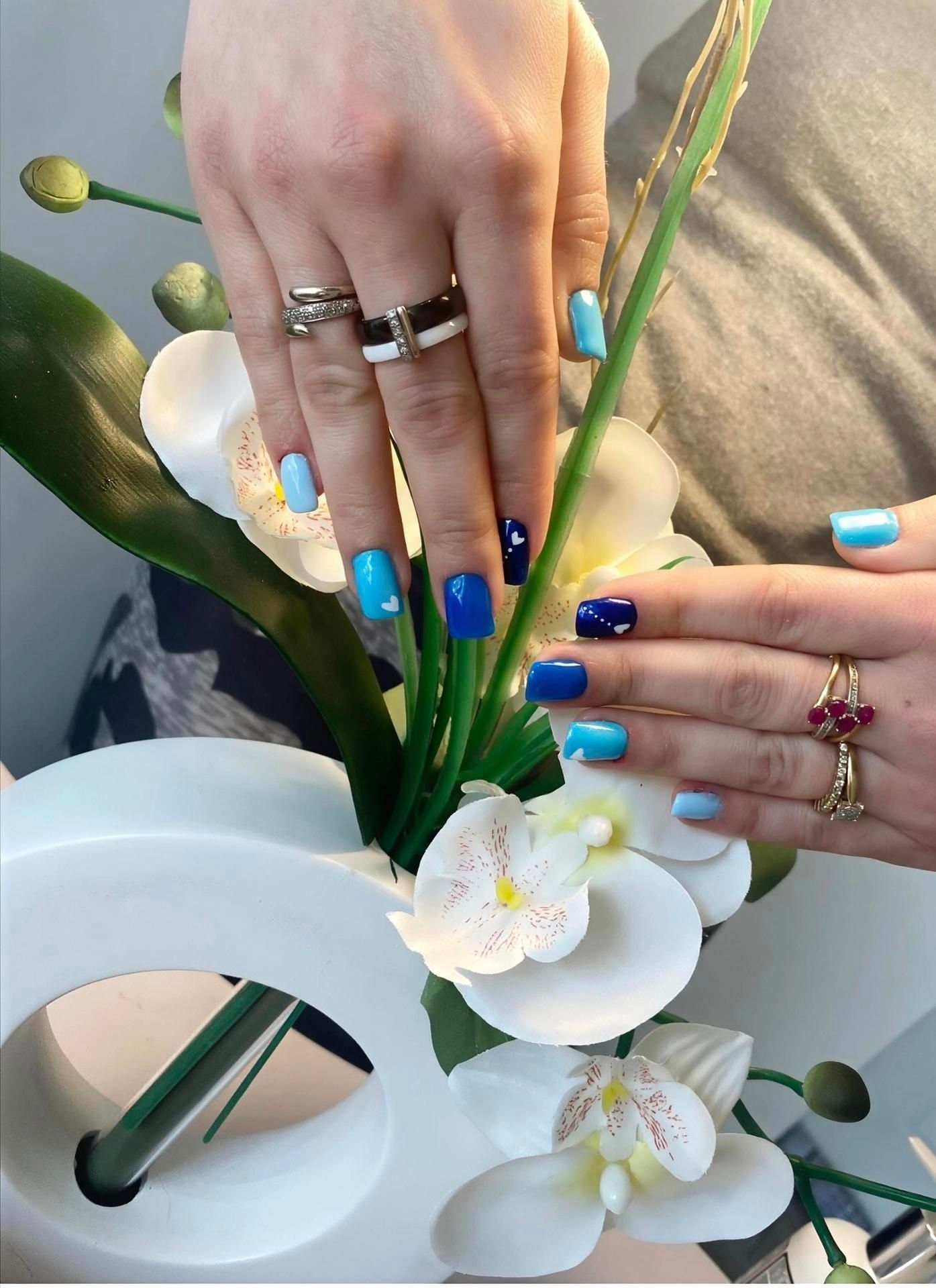 Des mains arrangeant des fleurs blanches dans un vase, avec des ongles peints en bleu et des bagues en argent.