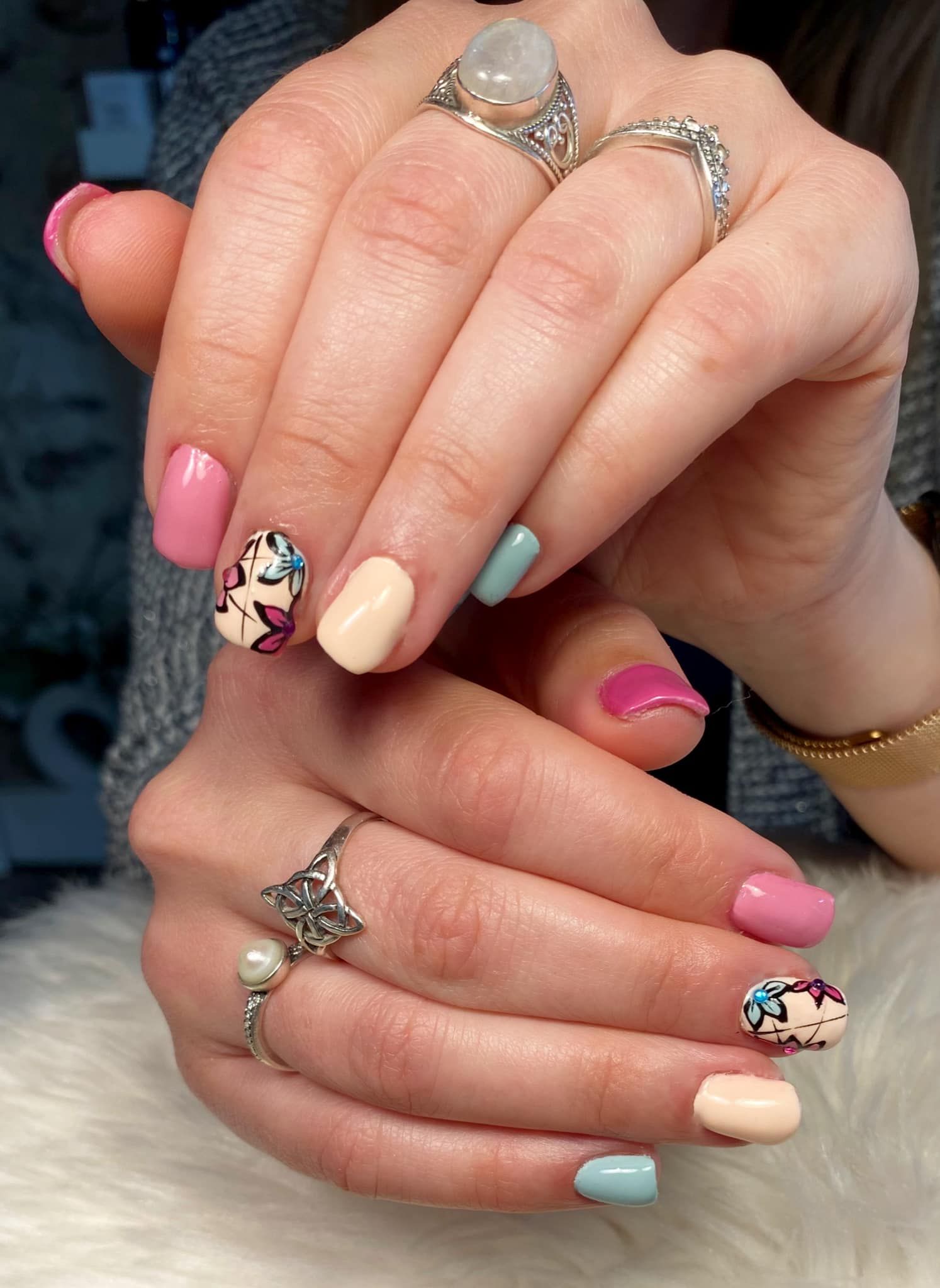 Mains aux ongles décorés de motifs pastel et ornées de bagues argentées, jointes sur un fond sombre.