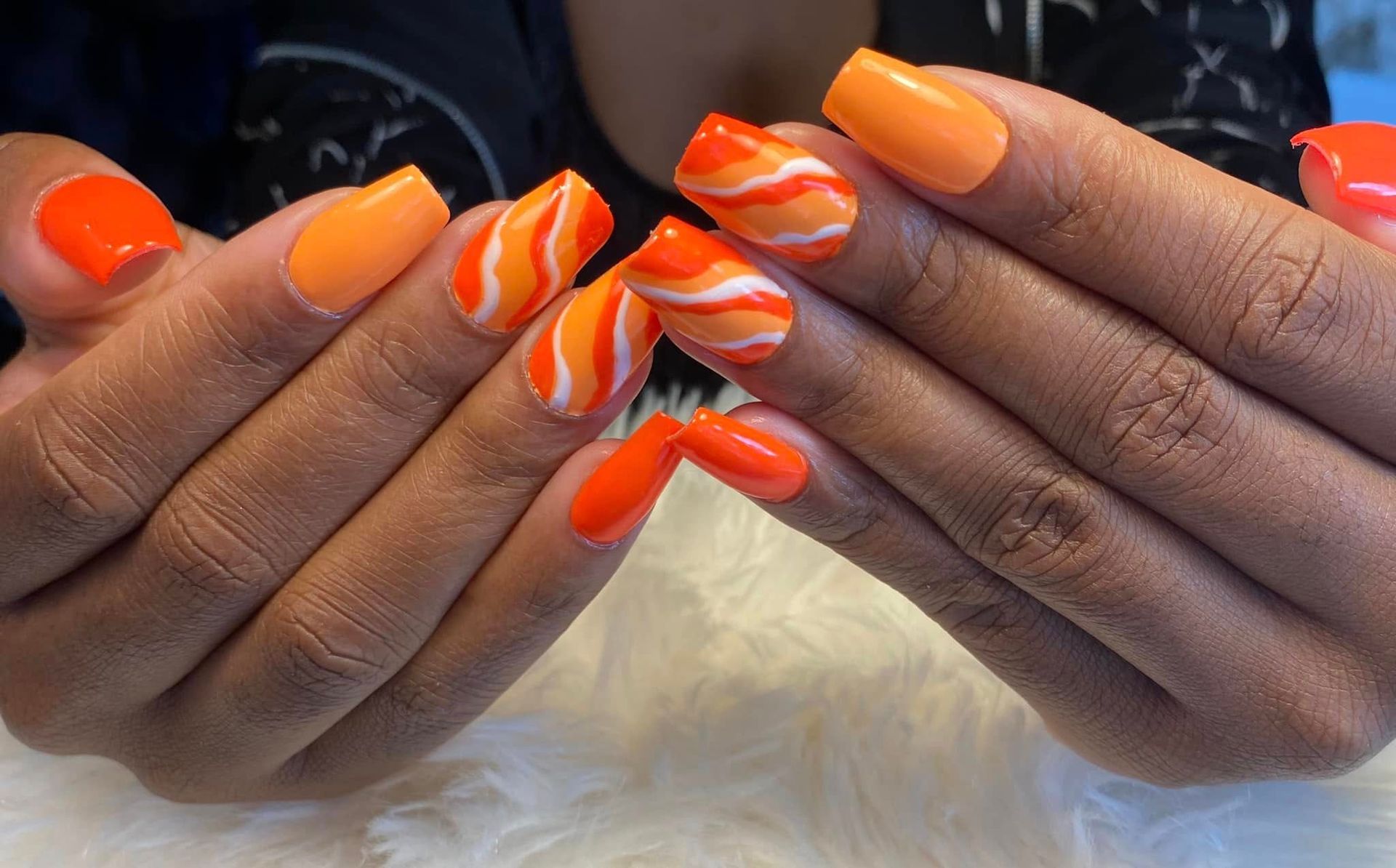 Des mains aux ongles manucurés, ornés de spirales orange vif et blanches, sur une surface blanche et duveteuse.
