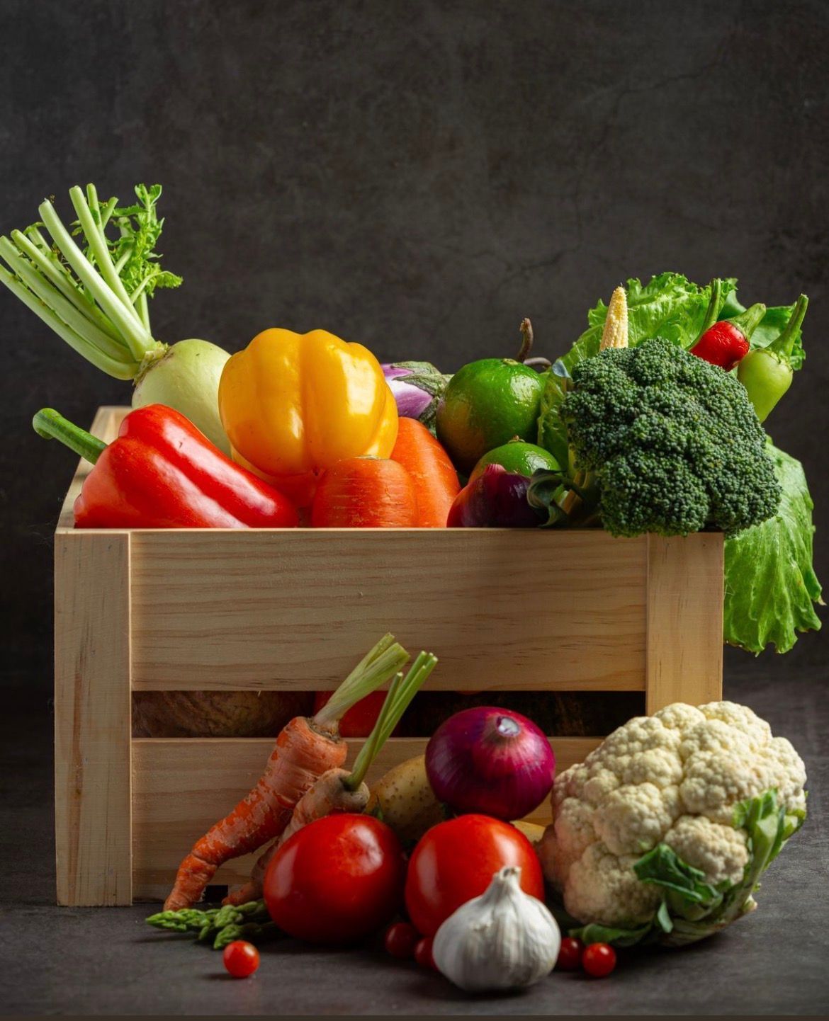 Légumes frais variés dans une caisse en bois sur fond sombre : poivrons, brocolis, tomates, ail et chou-fleur