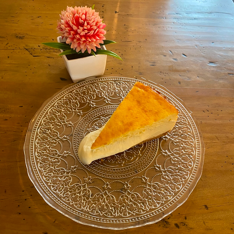 Una rebanada de tarta de queso está en un plato de vidrio sobre una mesa de madera.
