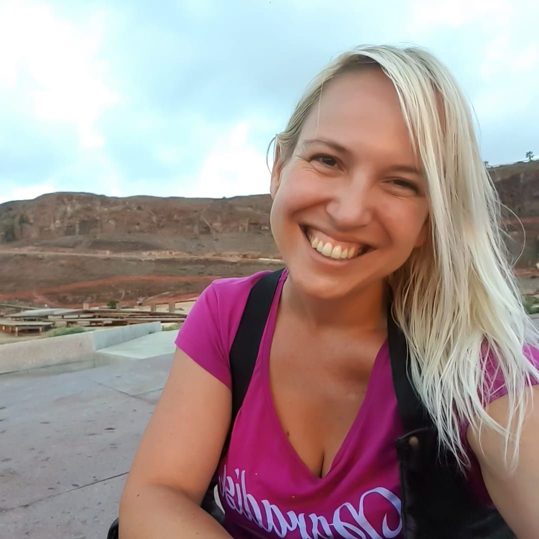 Mujer rubia sonriendo, vistiendo una camisa rosa, al aire libre con un fondo montañoso.