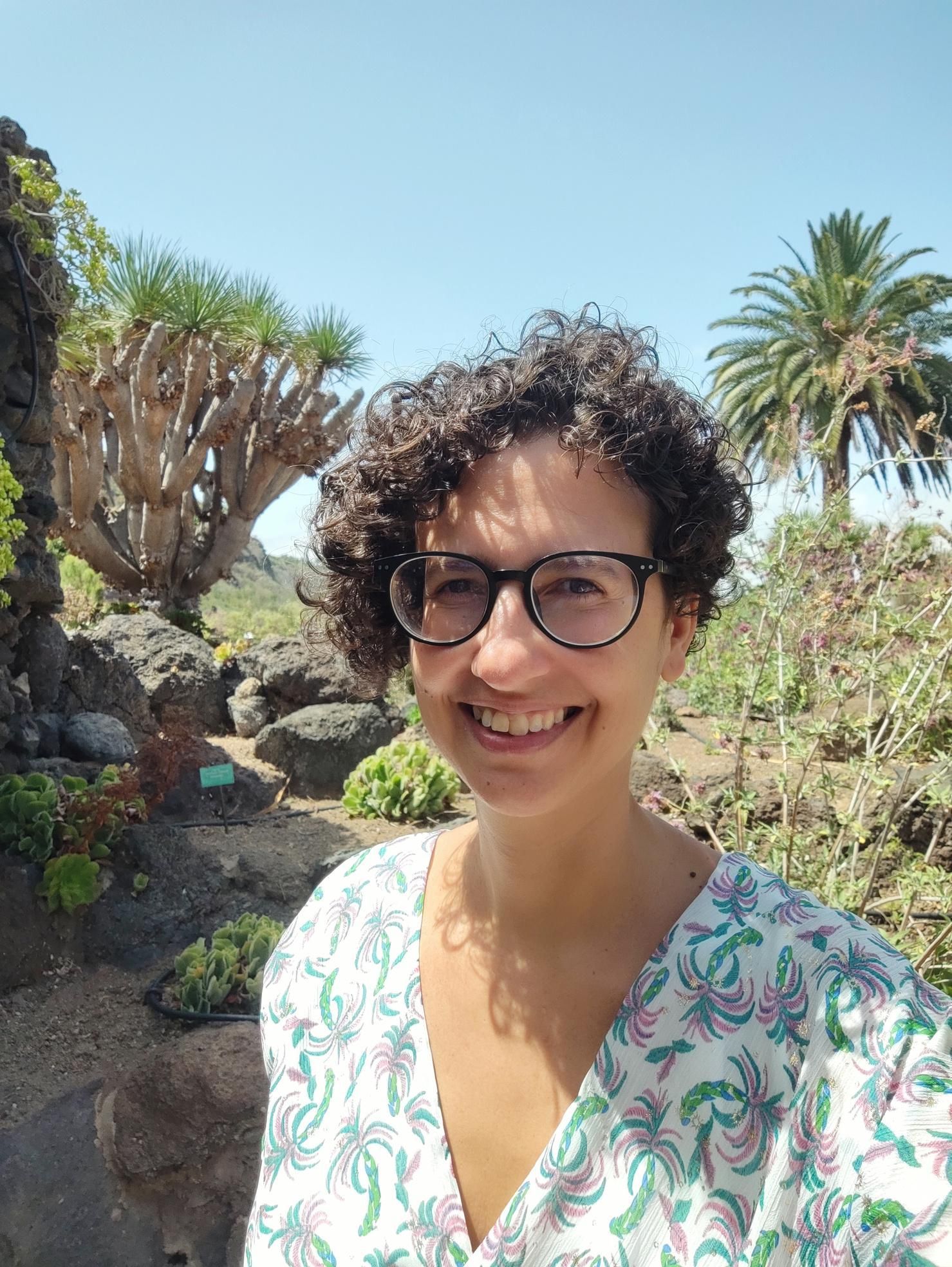 Mujer con gafas sonriendo en un jardín botánico, con árboles únicos y una palmera bajo un cielo azul.