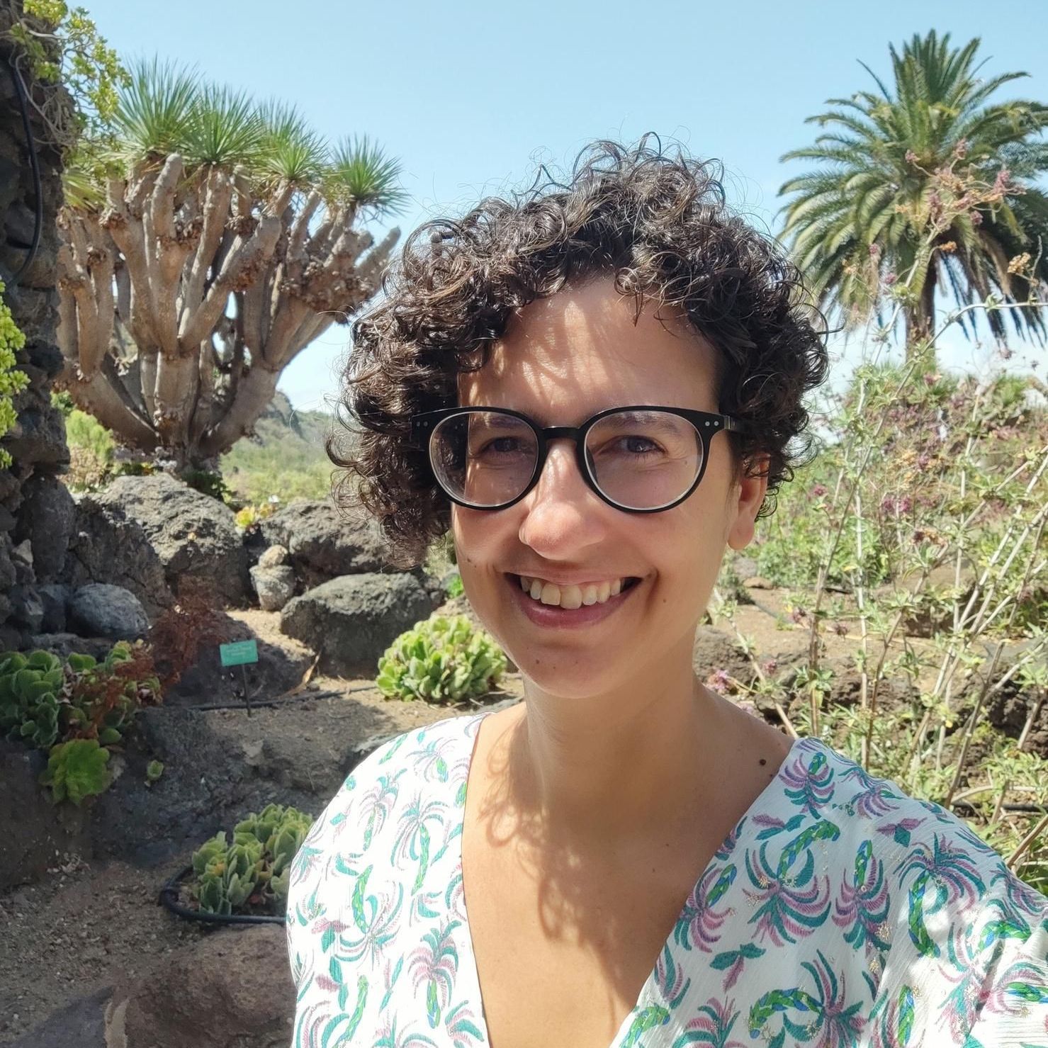 Mujer con cabello rizado y gafas sonriendo en un jardín botánico con cactus y palmeras.