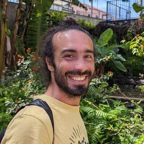 Hombre con barba, sonriendo, con camisa amarilla, al aire libre en medio de la vegetación.