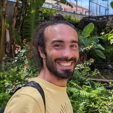 Hombre con barba sonriendo frente a la vegetación. Lleva una camisa amarilla y el pelo recogido en un moño.
