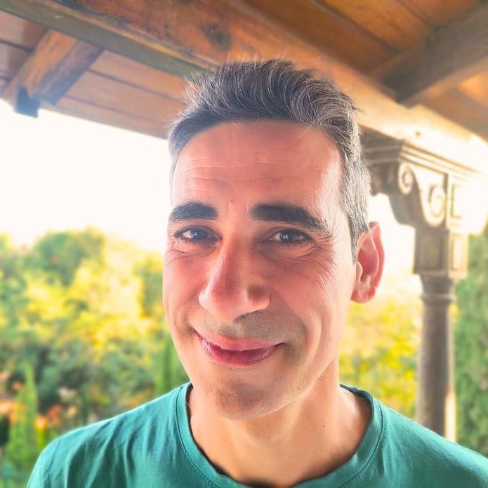 Hombre de pelo corto y oscuro sonriendo al aire libre, con camisa verde. Árboles y columnas desenfocados al fondo.