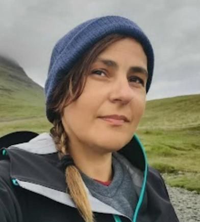 Mujer con cabello castaño, con gorro azul y trenza, chaqueta oscura, sonriendo levemente, al aire libre cerca de una montaña.