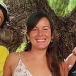 Mujer sonriendo, vistiendo un top de tirantes finos con flores blancas, con cabello castaño, al aire libre.