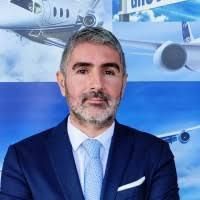 Hombre de traje azul, sonriendo, frente a aviones, posiblemente un ejecutivo de aviación.