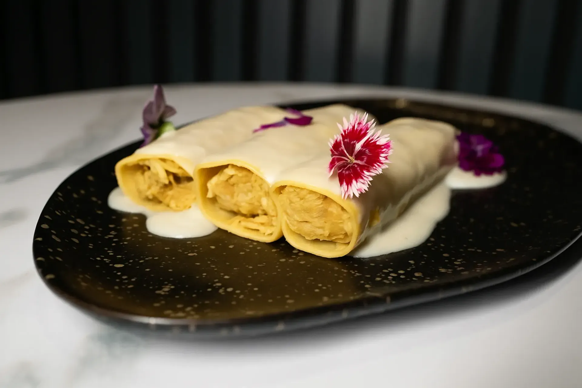 Tres canelones con salsa blanca y flores comestibles sobre plato negro.
