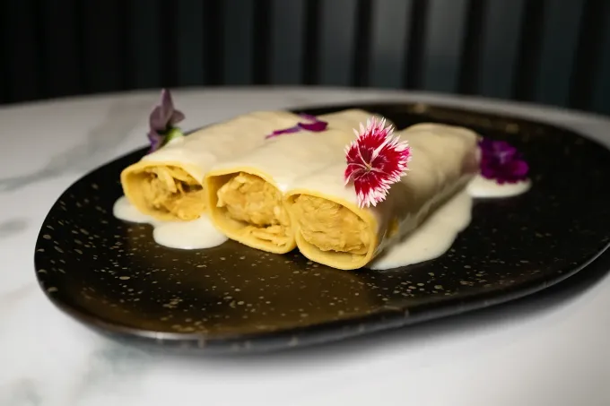 Tres canelones con salsa blanca y flores comestibles sobre plato negro.
