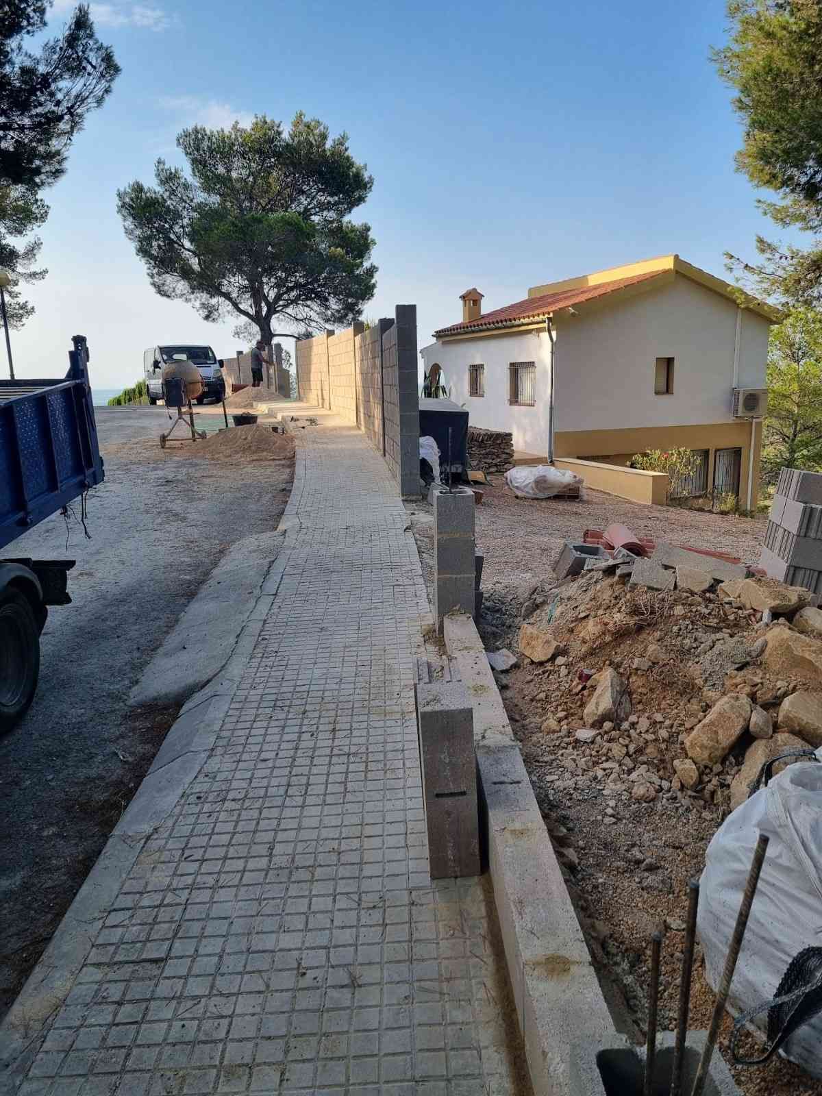 Sendero de piedra junto a una obra en construcción, con un camión azul, árboles y una casa blanca en la ladera de una colina.