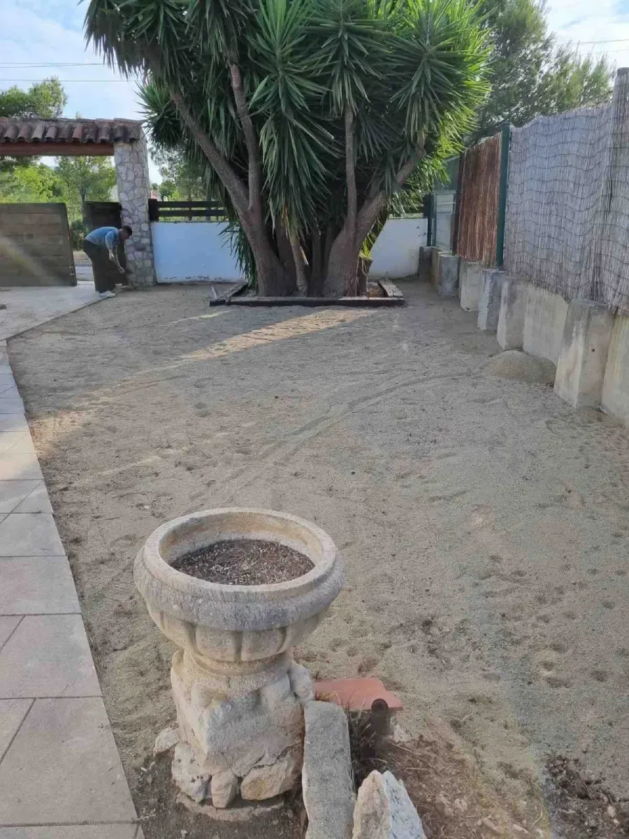 Patio trasero con una jardinera de piedra en primer plano, suelo de grava y un árbol junto a un muro y una puerta.