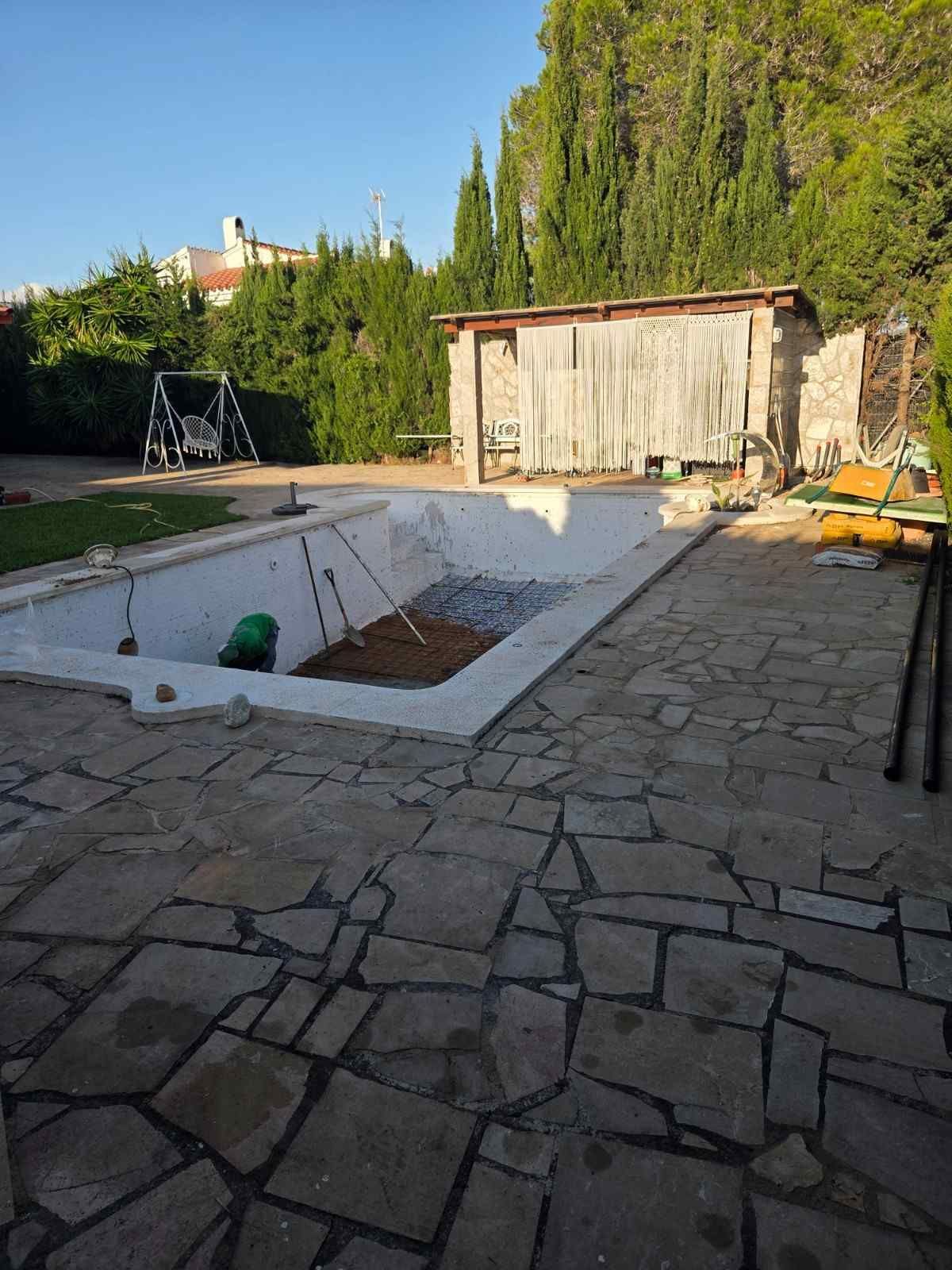 Piscina vacía en el patio trasero, junto a un patio de piedra, con un pequeño cobertizo y árboles altos al fondo.