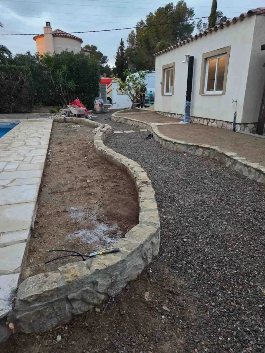 Sendero curvo de grava junto a una casa blanca y una jardinera de piedra en un patio en construcción.