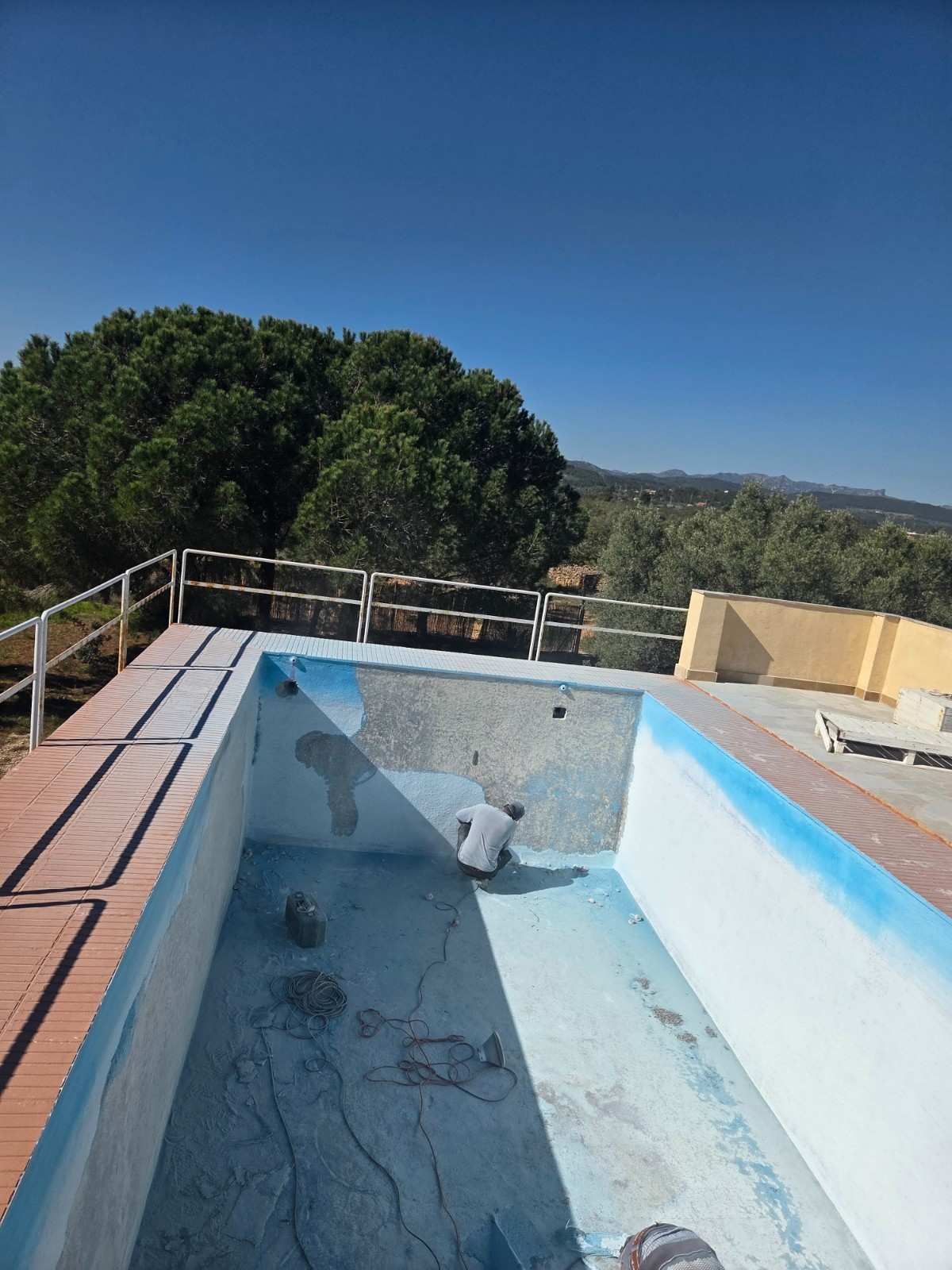 Piscina vacía junto a una terraza, con paredes pintadas de azul y árboles más allá de la barandilla.