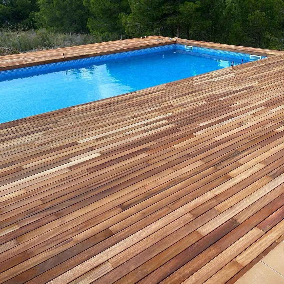 Terraza de madera junto a una piscina de color azul brillante, con árboles verdes al fondo.