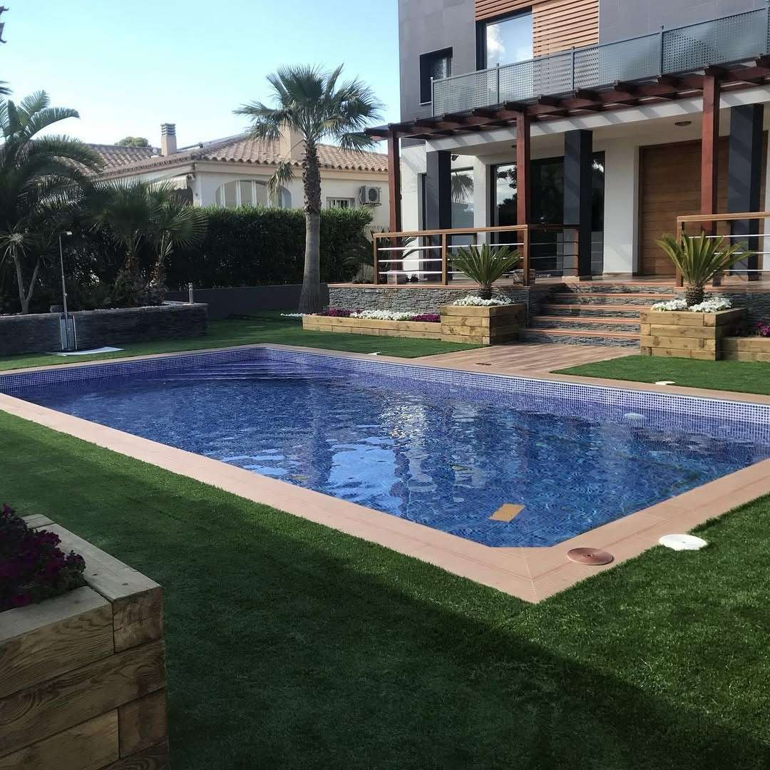 Patio trasero de una casa moderna con piscina de azulejos azules, césped y patio con escalones y palmeras.