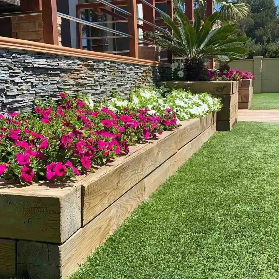 Parterre elevado de madera con flores rosas y blancas junto a un césped bien cuidado y un muro de piedra.