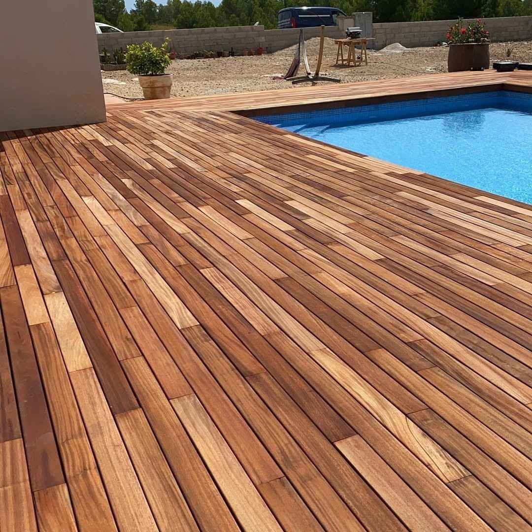 Terraza de madera junto a una piscina azul en un patio soleado al aire libre.