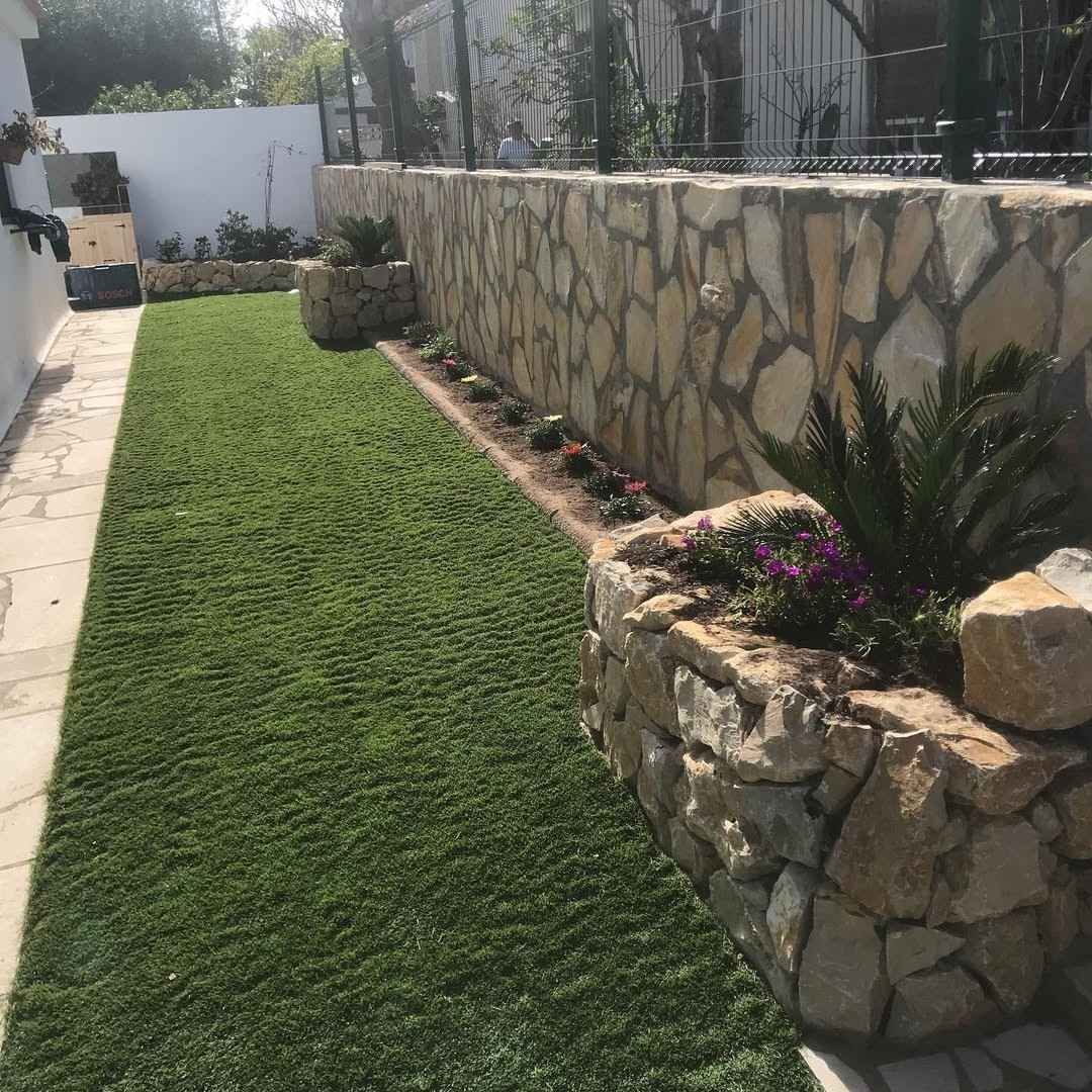 Muro de piedra ajardinado junto a un césped verde con pequeños macizos de flores bañados por la luz del sol.