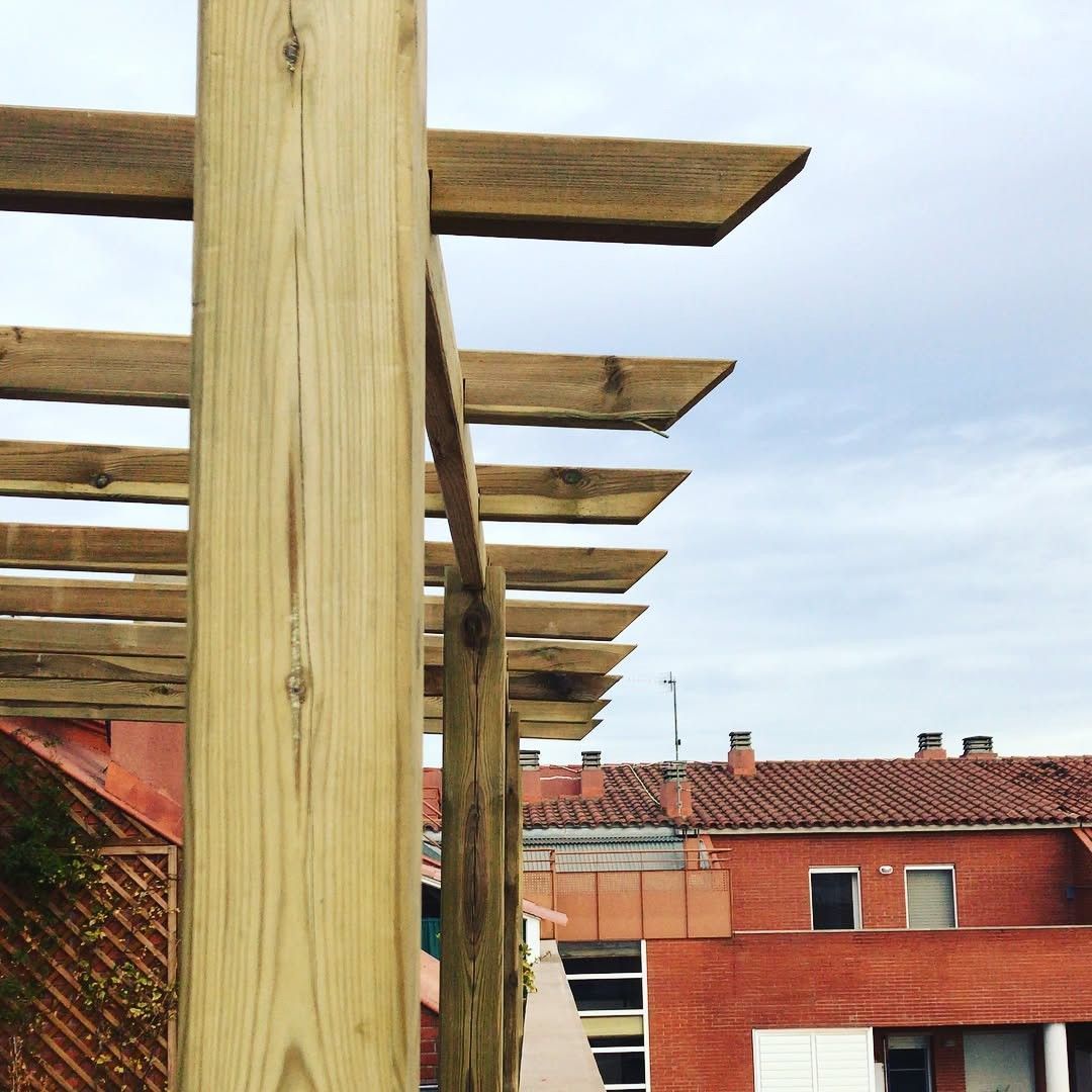 Vigas de pérgola de madera junto a edificios de tejados rojos bajo un cielo nublado.