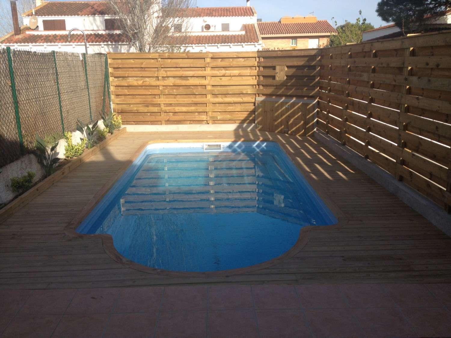 Pequeña piscina en el patio trasero con agua azul, rodeada de una terraza de madera y una valla, bañada por el sol.